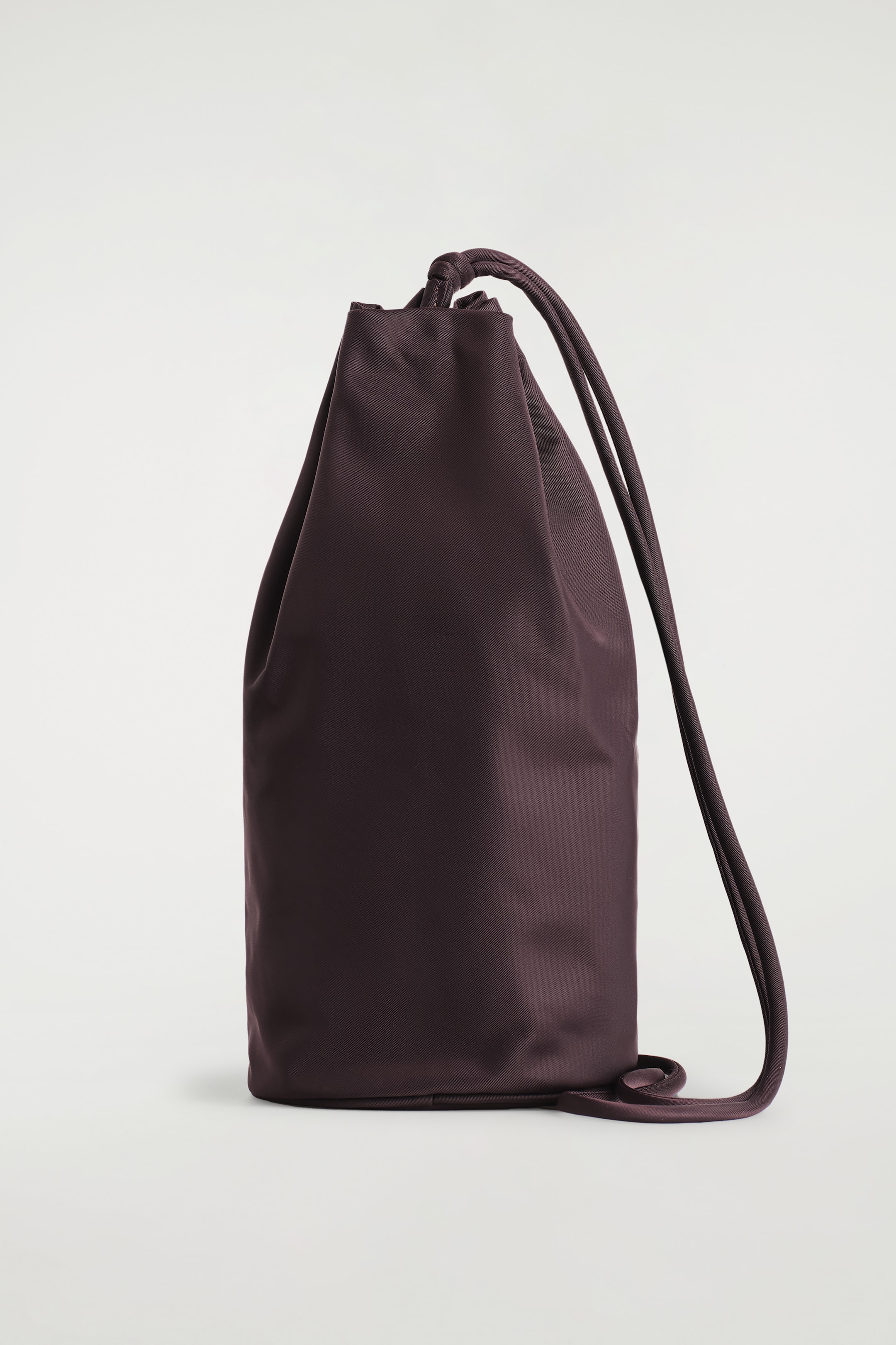 View larger image: A deep purple drawstring bag is presented from the front, featuring a soft, cylindrical body crafted from a smooth, slightly lustrous fabric. Its top is cinching closed, with dual drawstrings extending down the sides and pooling at the base.