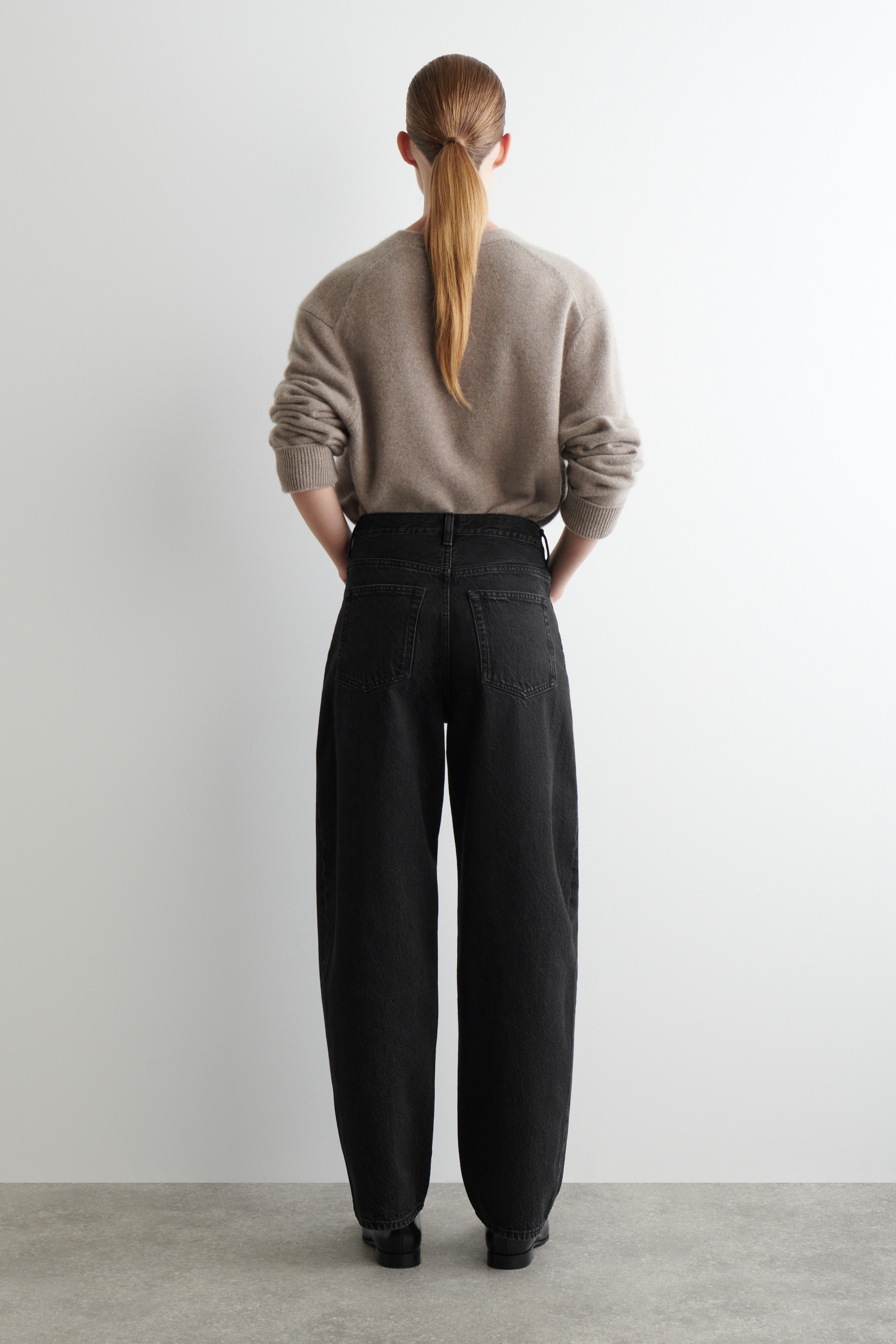 Visualizza immagine più grande: I jeans in denim nero sono presentati dal retro, mettendo in mostra la loro vestibilità a vita alta, a gamba larga e due tasche applicate visibili sul retro. Un maglione girocollo marrone chiaro con polsini a coste è indossato, appoggiandosi alla cintura dei jeans.