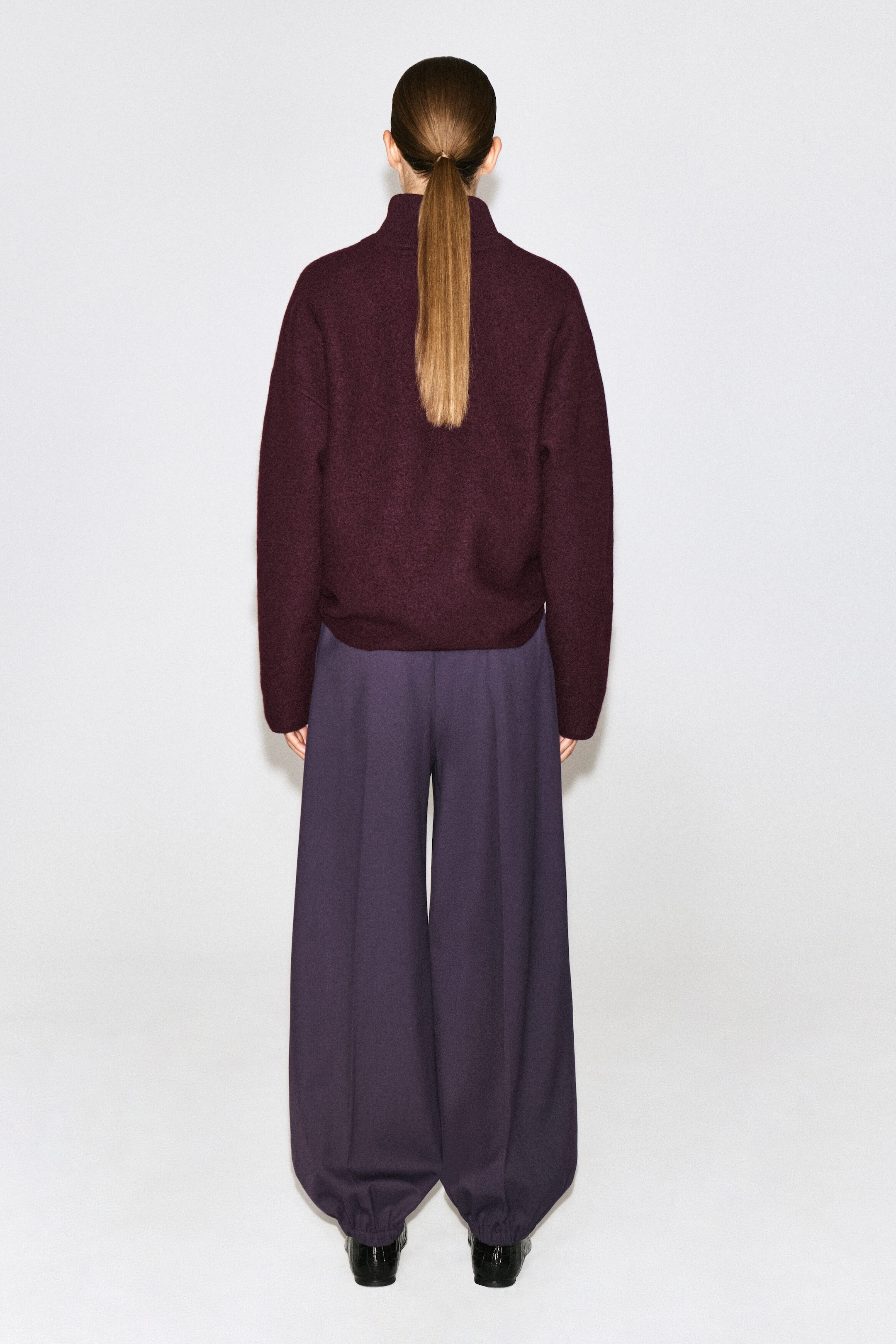Un chandail en laine bouillie bordeaux avec un col montant et une coupe décontractée est porté par le mannequin, vu de dos. Le chandail est agencé avec un pantalon ample pourvu de jambes larges, de couleur violet foncé. Un chandail en laine bouillie bordeaux avec un col montant et une coupe décontractée est porté par le mannequin, vu de dos. Le chandail est agencé avec un pantalon ample pourvu de jambes larges, de couleur violet foncé.