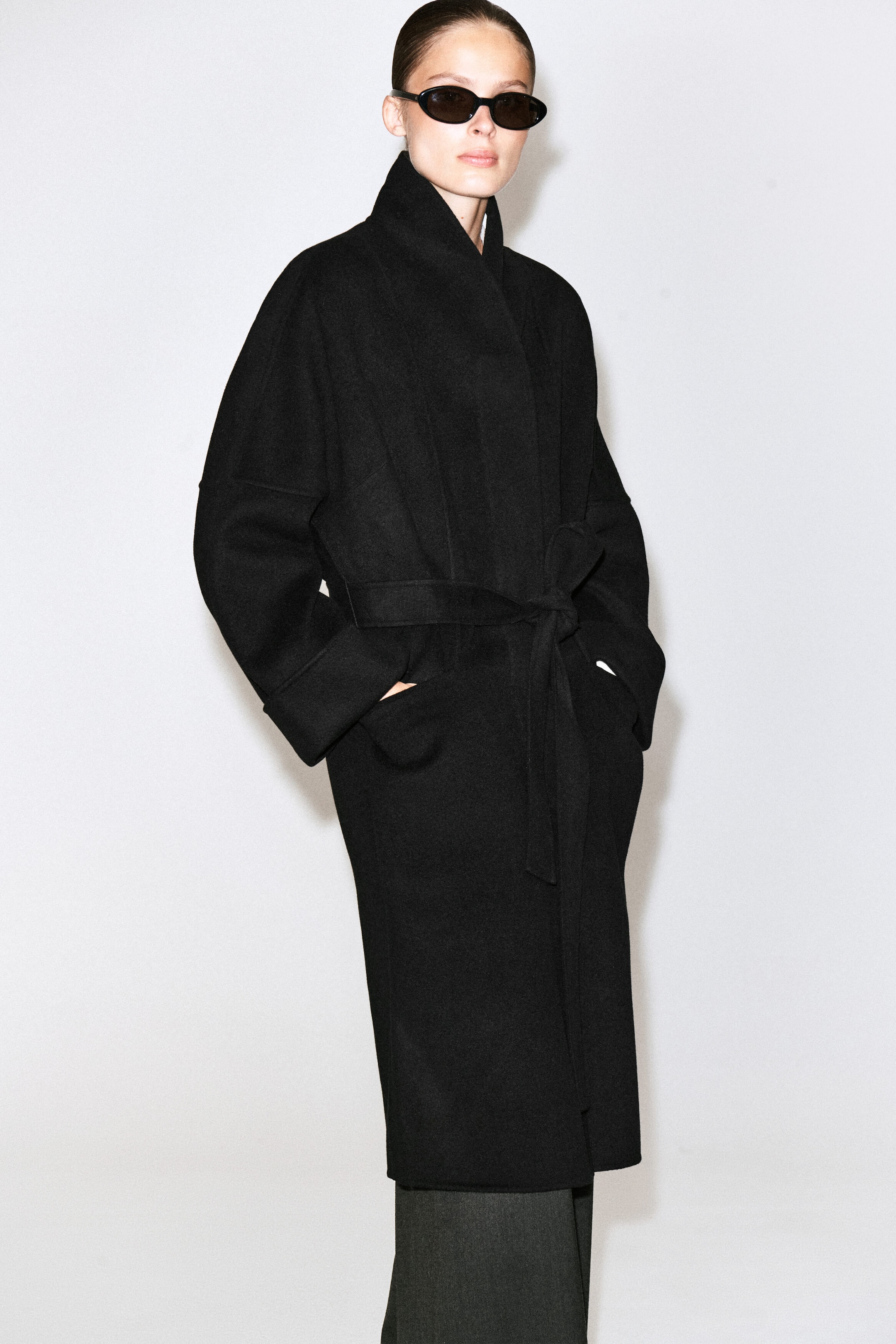 Le manteau châle oversize en laine double face noir est porté par le mannequin, qui se tient debout, les mains glissées dans les poches latérales. Le manteau présente un grand col châle structuré, des épaules tombantes et une ceinture assortie nouée à la taille. Le manteau châle oversize en laine double face noir est porté par le mannequin, qui se tient debout, les mains glissées dans les poches latérales. Le manteau présente un grand col châle structuré, des épaules tombantes et une ceinture assortie nouée à la taille.