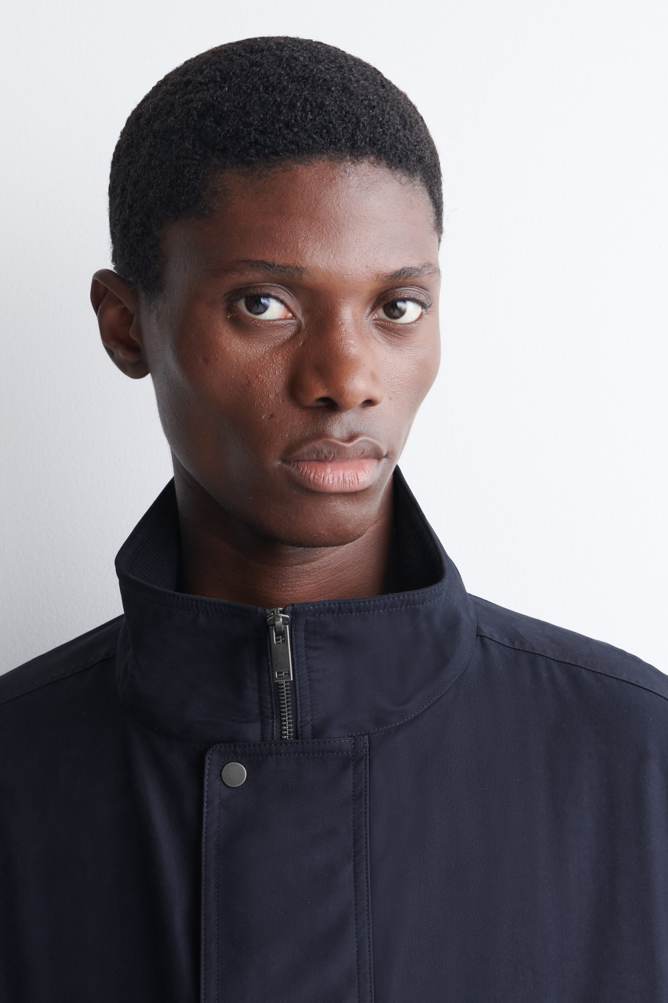 Une veste bomber rembourrée à col montant, d’un bleu marine foncé, est portée par le mannequin. La veste présente un col montant avec une fermeture éclair avant partiellement fermée et un rabat pare-tempête doté d’un bouton-pression métallique visible.