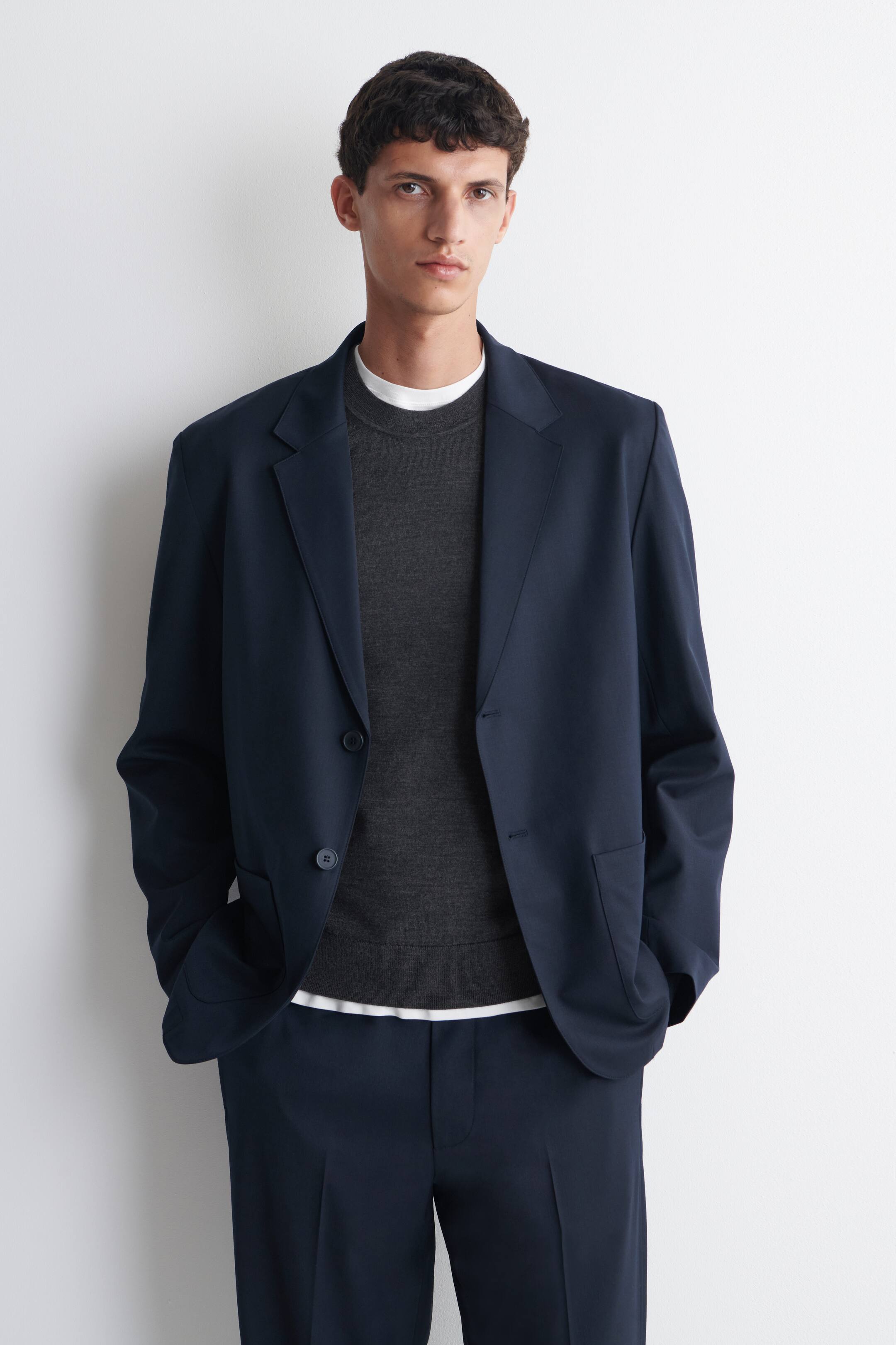 Agrandir l'image: Le mannequin porte un blazer bleu marine déstructuré, doté de revers crantés et de poches plaquées à l’avant. Le blazer est porté ouvert sur un pull gris à col rond et un t-shirt blanc, et associé à un pantalon bleu marine assorti, tandis que le mannequin glisse les mains dans les poches du blazer.