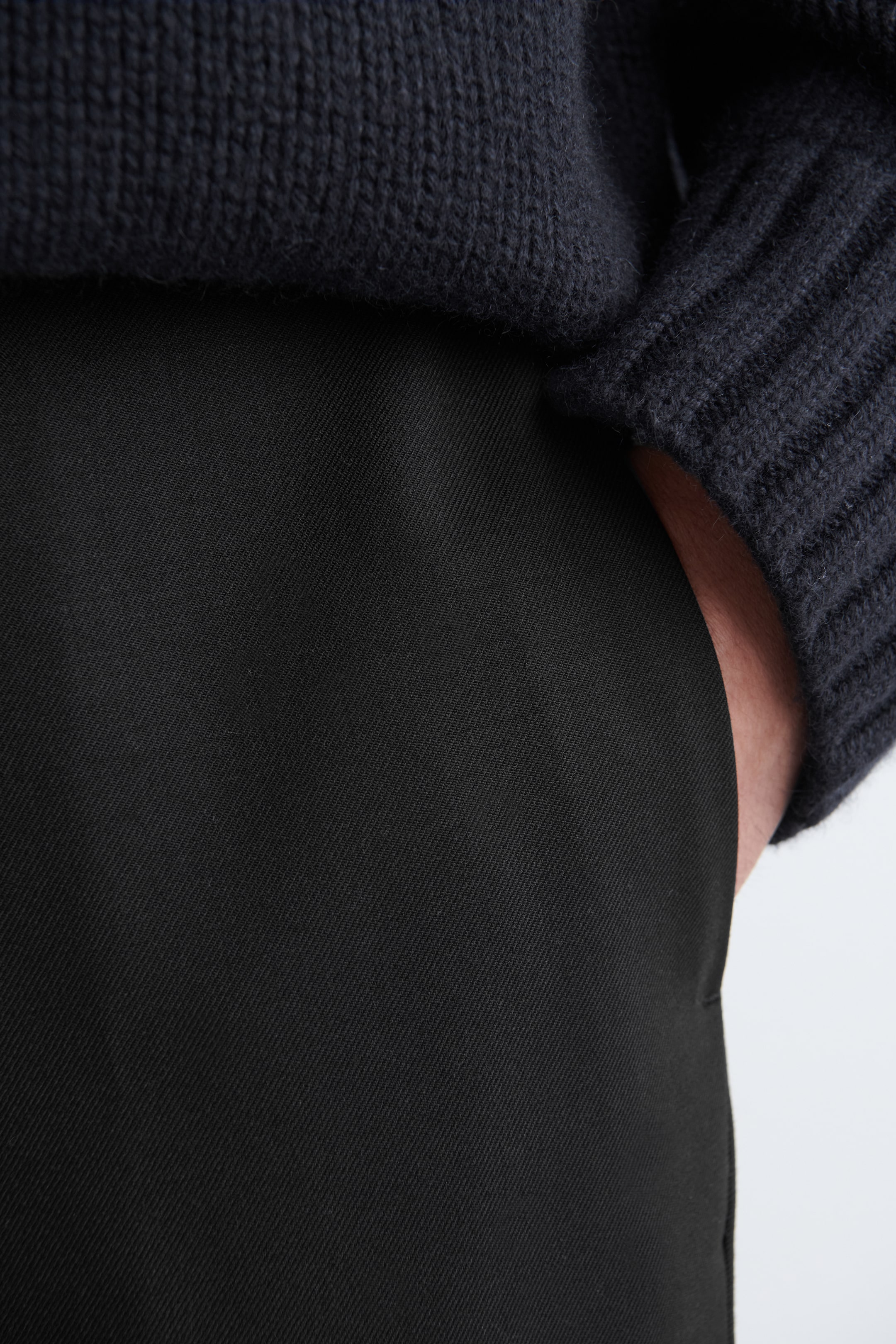 Visualizza immagine più grande: I pantaloni neri sono mostrati da una vista laterale-frontale, caratterizzati da un sottile tessuto a trama diagonale. Un maglione scuro con un colletto a coste sovrapposto alla cintura, e una mano è appoggiata nella tasca laterale dei pantaloni.