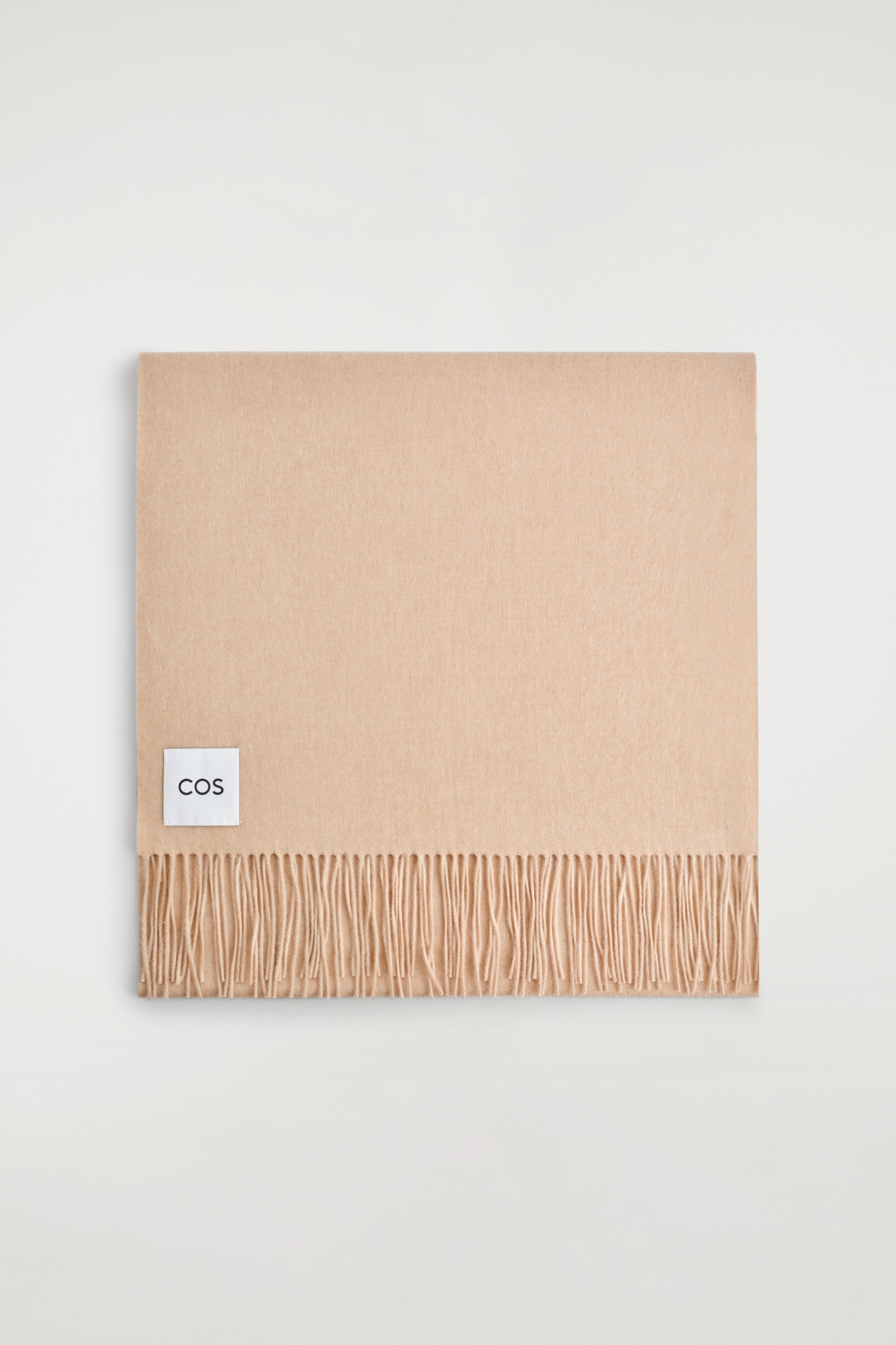 Visualizza immagine più grande: Una sciarpa larga di colore beige chiaro è distesa piatta, vista dal davanti. Presenta una texture liscia, frange lungo un'estremità e un'etichetta rettangolare bianca con la scritta "COS" cucita sul lato sinistro.