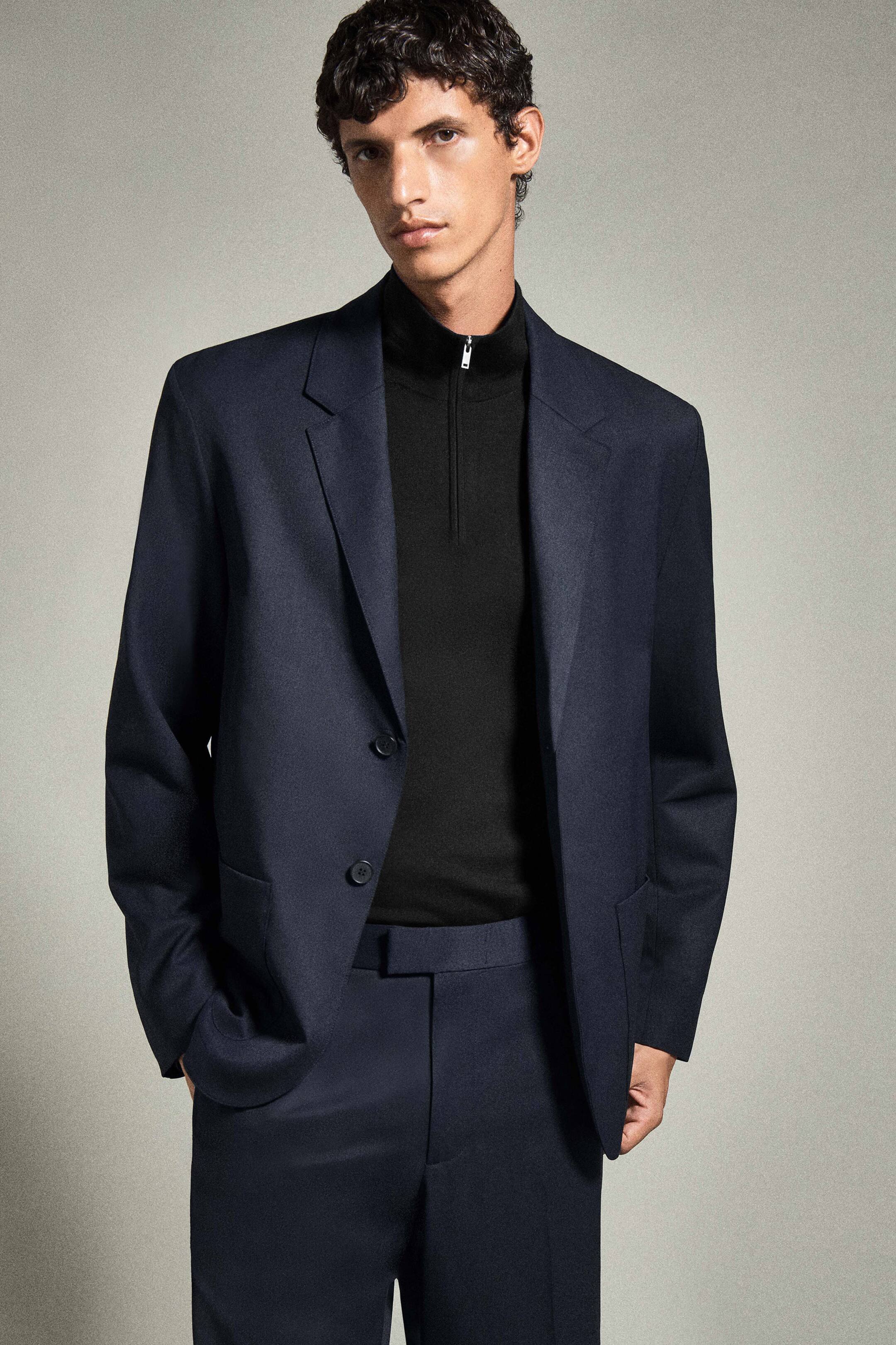 Visualizza l'immagine ingrandita: Il modello indossa frontalmente un blazer blu navy anti piega, caratterizzato da revers a lancia e una struttura rilassata. Indossa il blazer sopra un pullover nero in lana merino con collo a lupetto e zip, dotato di chiusura a quarto di zip color argento.