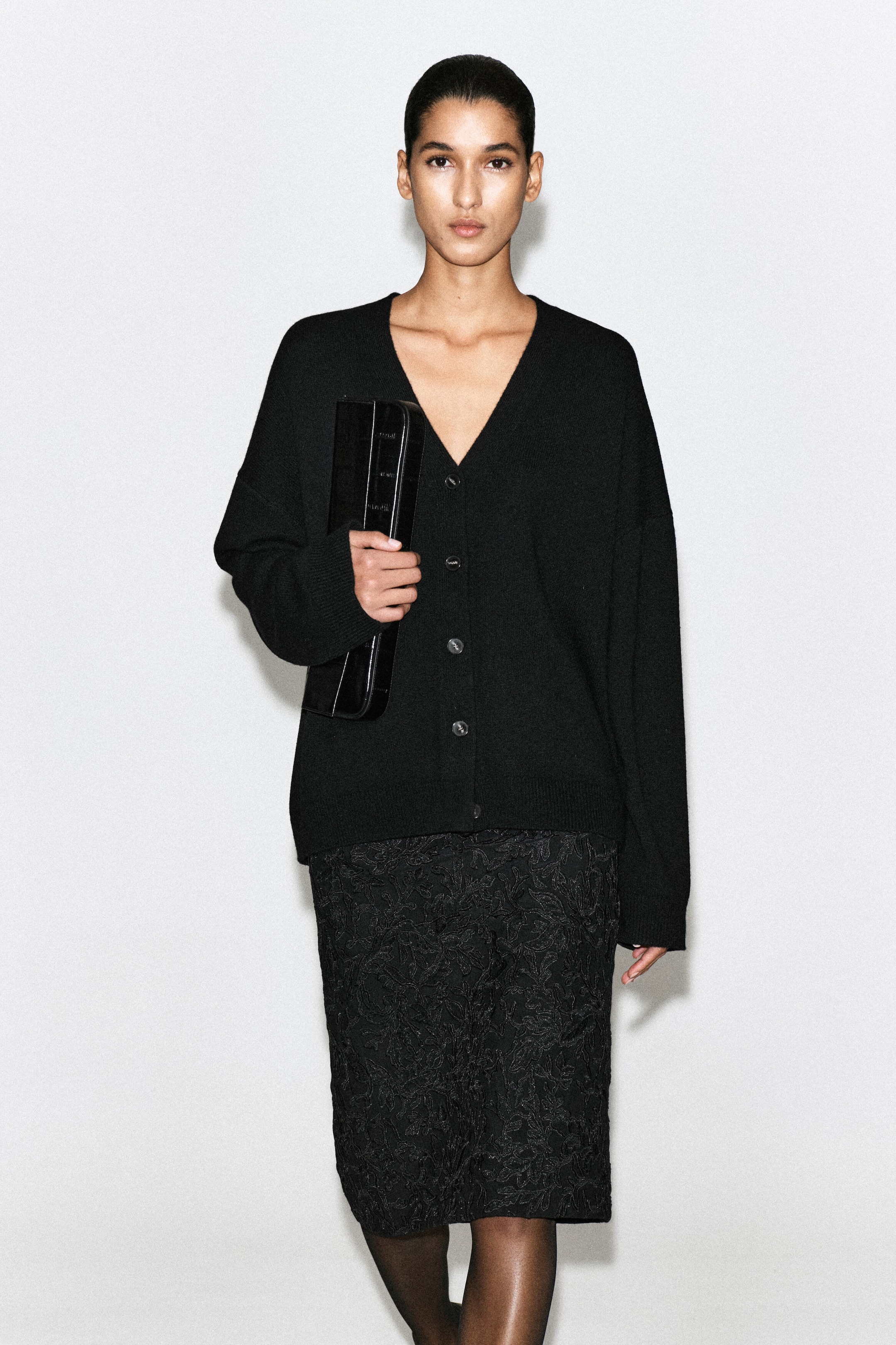 Un cardigan nero a V dalla vestibilità morbida viene indossato frontalmente, con spalle scese, chiusura con bottoni e finiture a coste. Il cardigan è abbinato a una gonna nera texturizzata.
