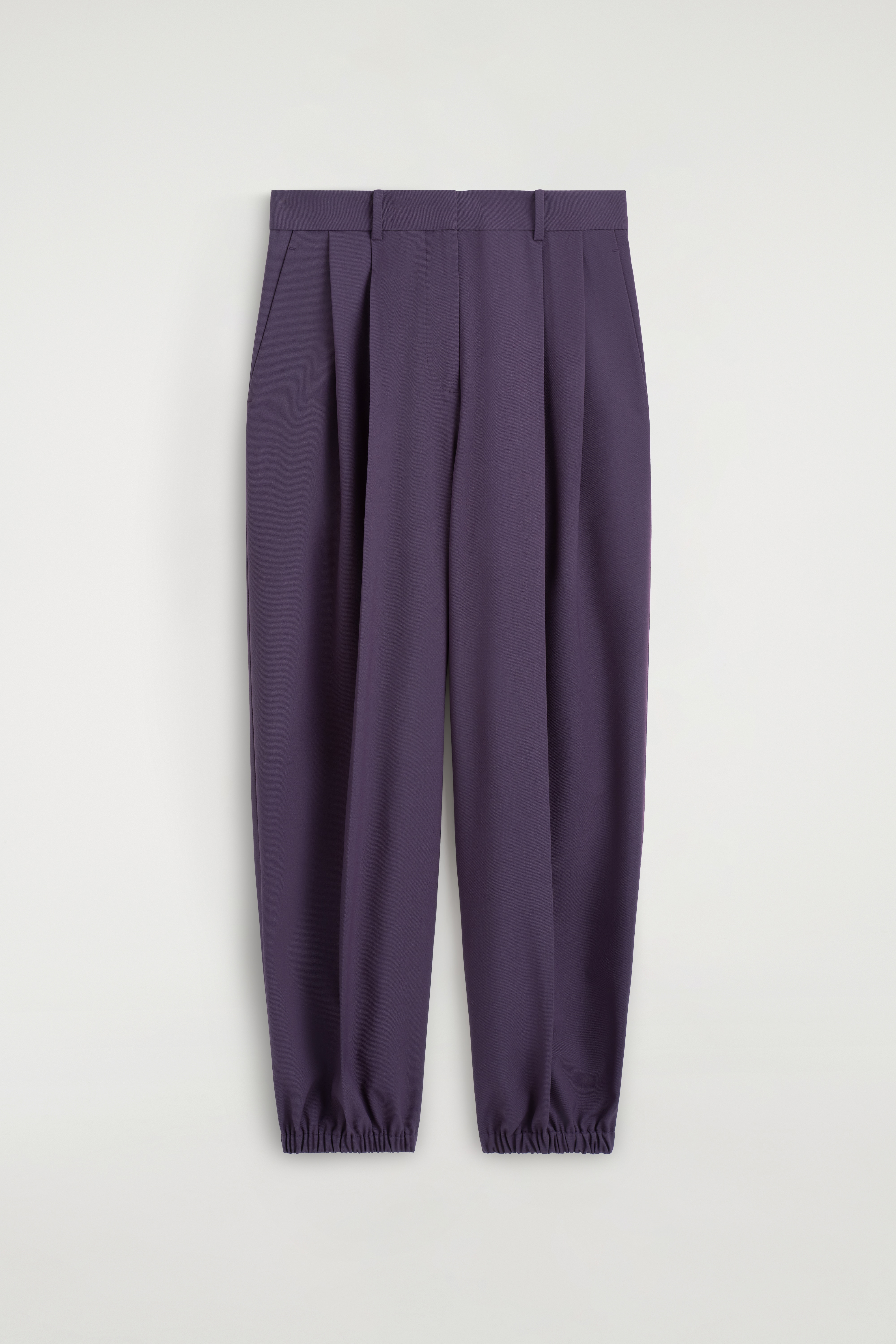 Le mannequin porte un pantalon ballon plissé dans une teinte violet foncé, doté dâune taille haute avec des plis sur le devant et une jambe volumineuse resserrée à la cheville. Le pantalon est associé à un pull bordeaux rentré, à une ceinture marron foncé et à des mocassins en cuir noirs.