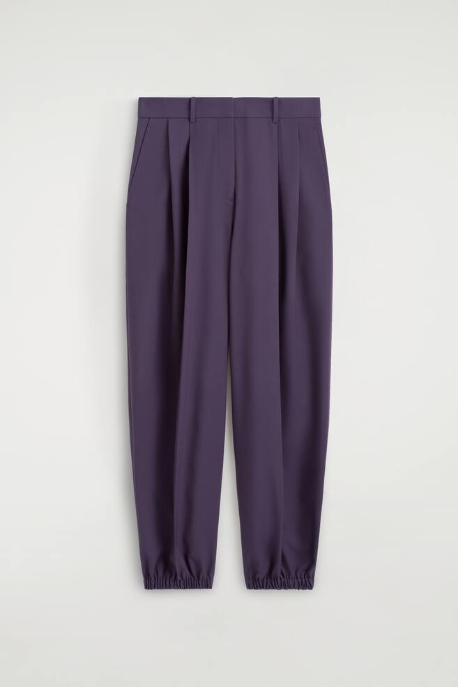 Pleated balloon trousers in a deep purple colour are shown from the front, featuring a high-waisted design with belt loops and deep front pleats that create a voluminous silhouette. The legs taper down to gathered, elasticated ankle cuffs. - 1