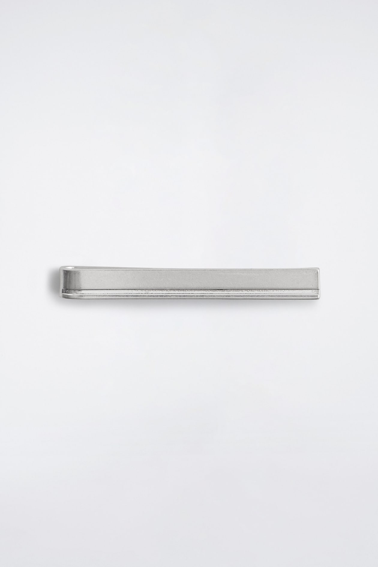 A silver-plated brass tie pin is laid horizontally from a front-facing perspective, showcasing its brushed matte finish and streamlined rectangular design. A subtle horizontal groove runs along the lower edge of the minimalist bar.