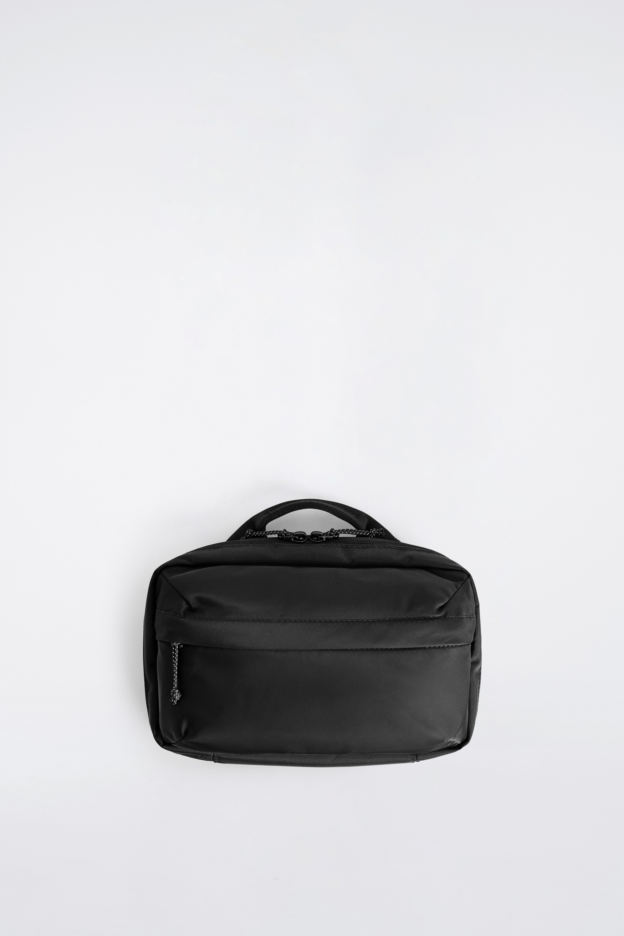 Agrandir l'image: Un sac rectangulaire noir est posé à plat, face vers l'avant, avec une poignée de transport sur le dessus et deux fermetures éclair principales avec des languettes de tirage en corde le long de son bord supérieur. Une poche avant horizontale zippée est également visible, accentuée par une languette de tirage en corde mouchetée sur la gauche.