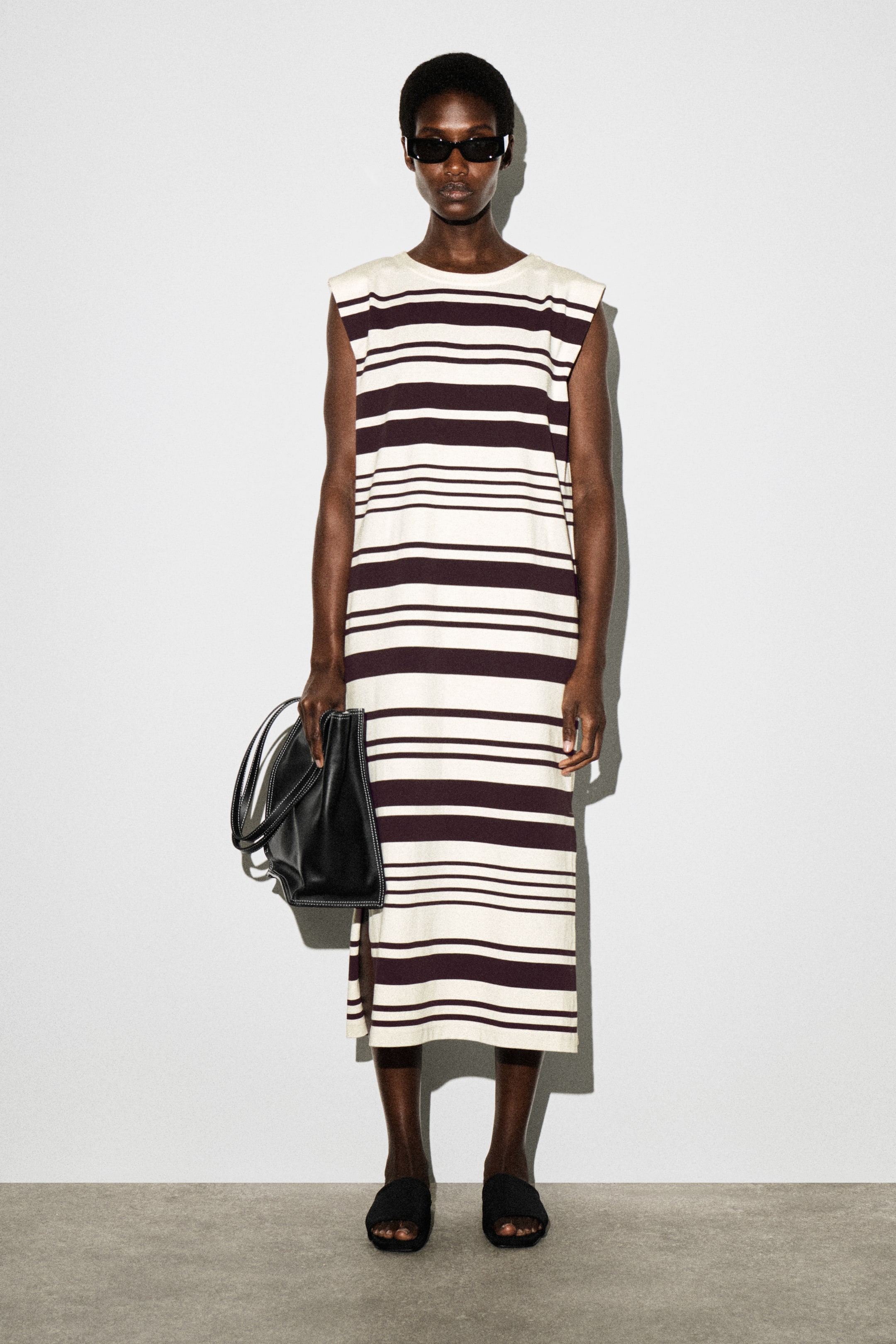 View larger image: The model is wearing a sleeveless midi dress with cream and dark brown horizontal stripes of varying widths. It features a crew neck and a side slit. The model is accessorising with black rectangular sunglasses, a black handbag, and black slide sandals.