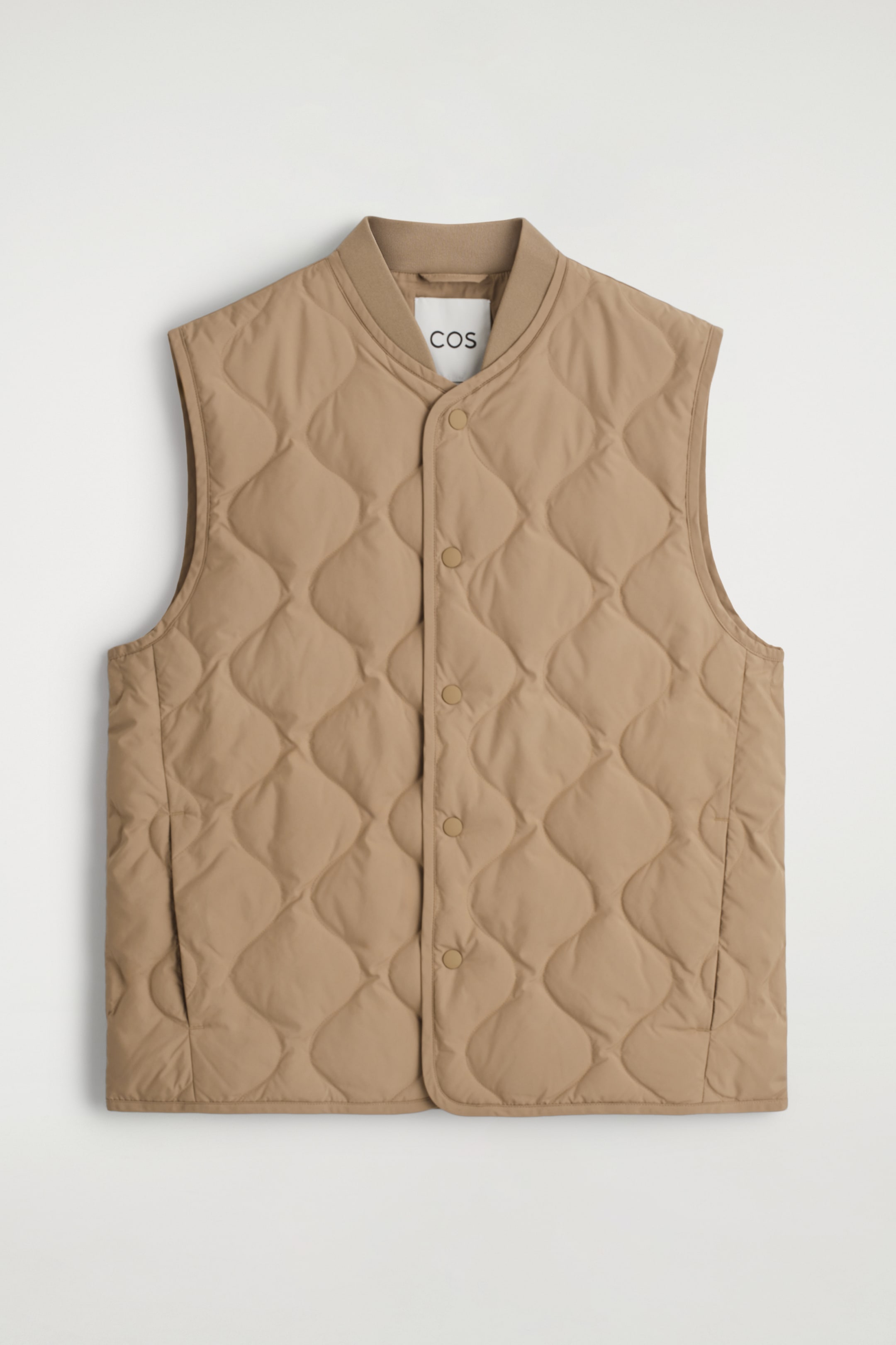 Agrandir l'image: Un gilet beige est exposé face à l'avant, avec un motif matelassé en losange sur tout le corps. Il est conçu avec un col montant côtelé et une ouverture frontale centrale sécurisée par des boutons-pression, avec deux poches passepoilées en biais également visibles sur le bas avant.