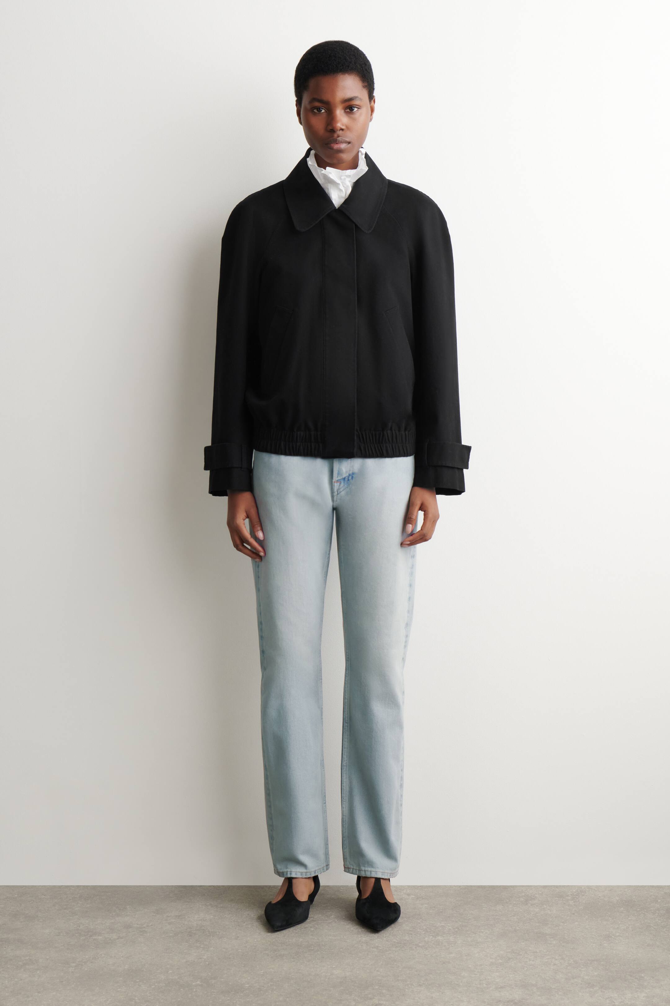 Agrandir l'image: La veste noire est dotée d'un col pointu classique et d'une fermeture frontale dissimulée. Elle est conçue avec une coupe décontractée, des manches raglan, des poignets larges et un ourlet élastiqué, avec un col de chemise blanc à volants visible en dessous.