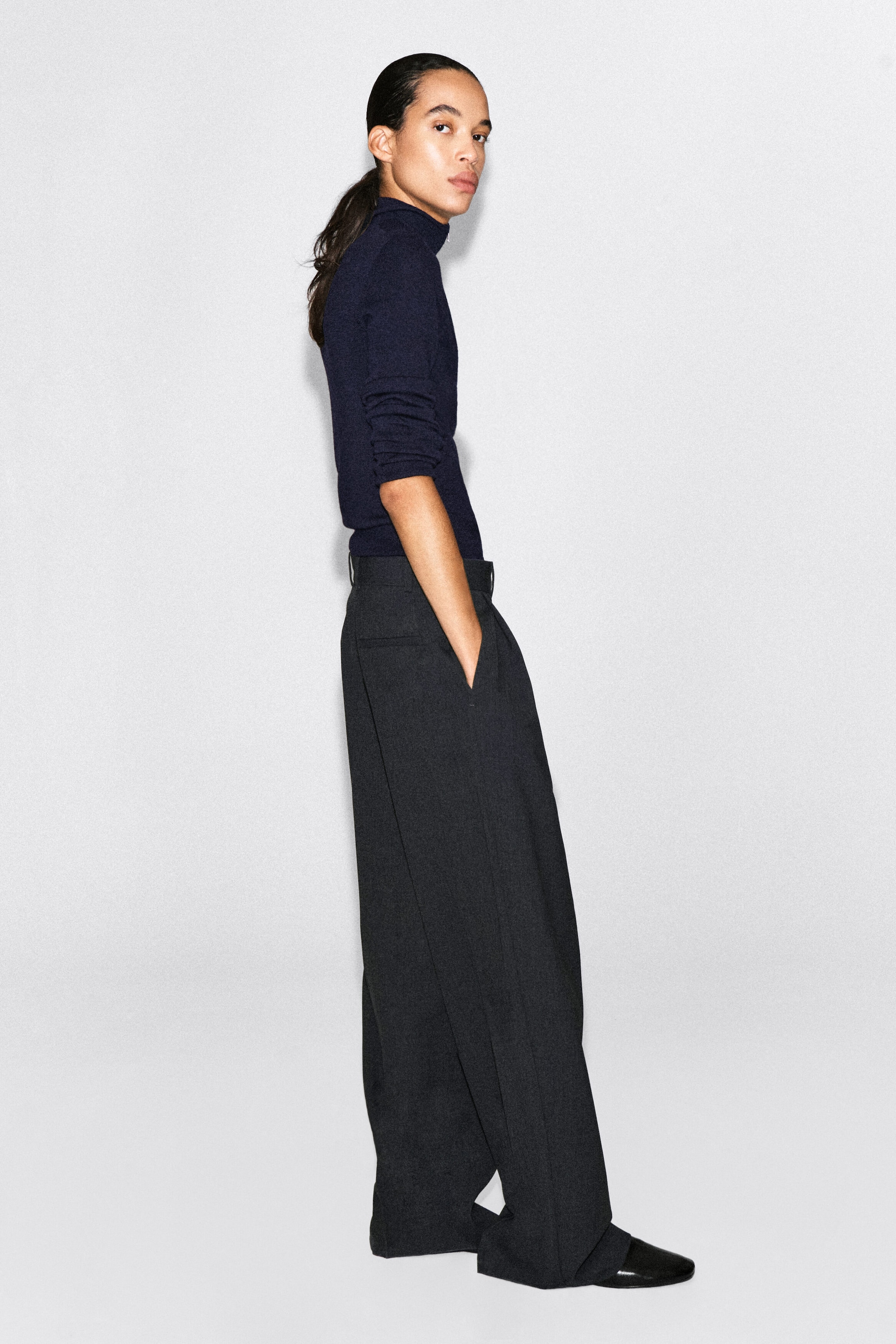 These high-waisted, wide-leg charcoal grey trousers are facing right, showcasing a relaxed, fluid drape and a side pocket where a hand is resting. The trousers are styled with a fitted navy blue turtleneck jumper and black pointed-toe slip-on shoes. These high-waisted, wide-leg charcoal grey trousers are facing right, showcasing a relaxed, fluid drape and a side pocket where a hand is resting. The trousers are styled with a fitted navy blue turtleneck jumper and black pointed-toe slip-on shoes.