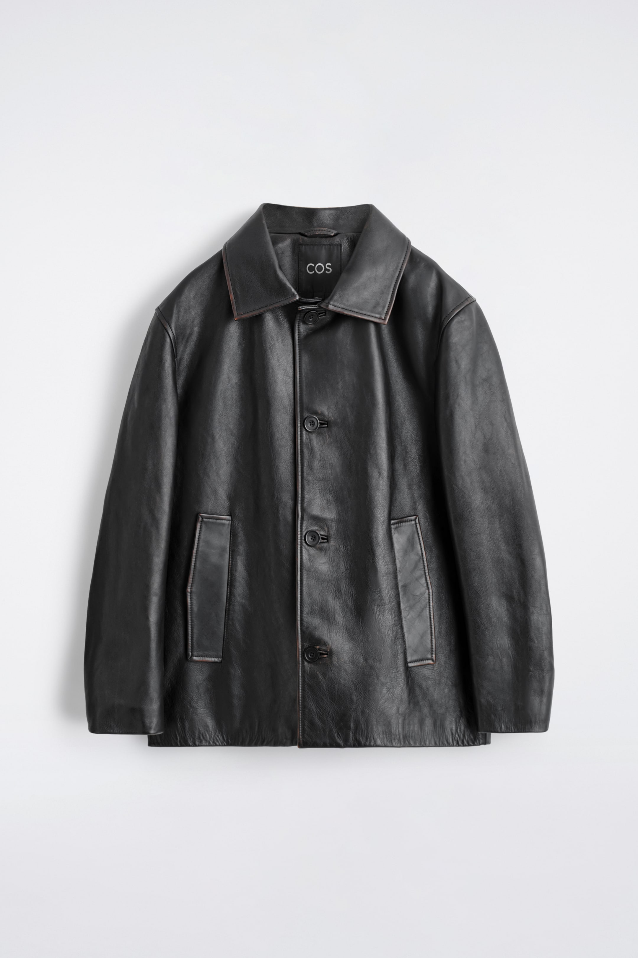 Agrandir l'image: Une veste en cuir noir à coupe décontractée est présentée de face, avec un col pointu classique doté d'une finition de bord visible de ton brun et d'une surpiqûre contrastante. Une étiquette de marque COS interne est également visible. Elle est conçue avec une fermeture à boutons sur le devant, mettant en valeur des boutons sombres, et deux grandes poches plaquées rectangulaires avec une finition de bord de ton brun assortie et une surpiqûre.