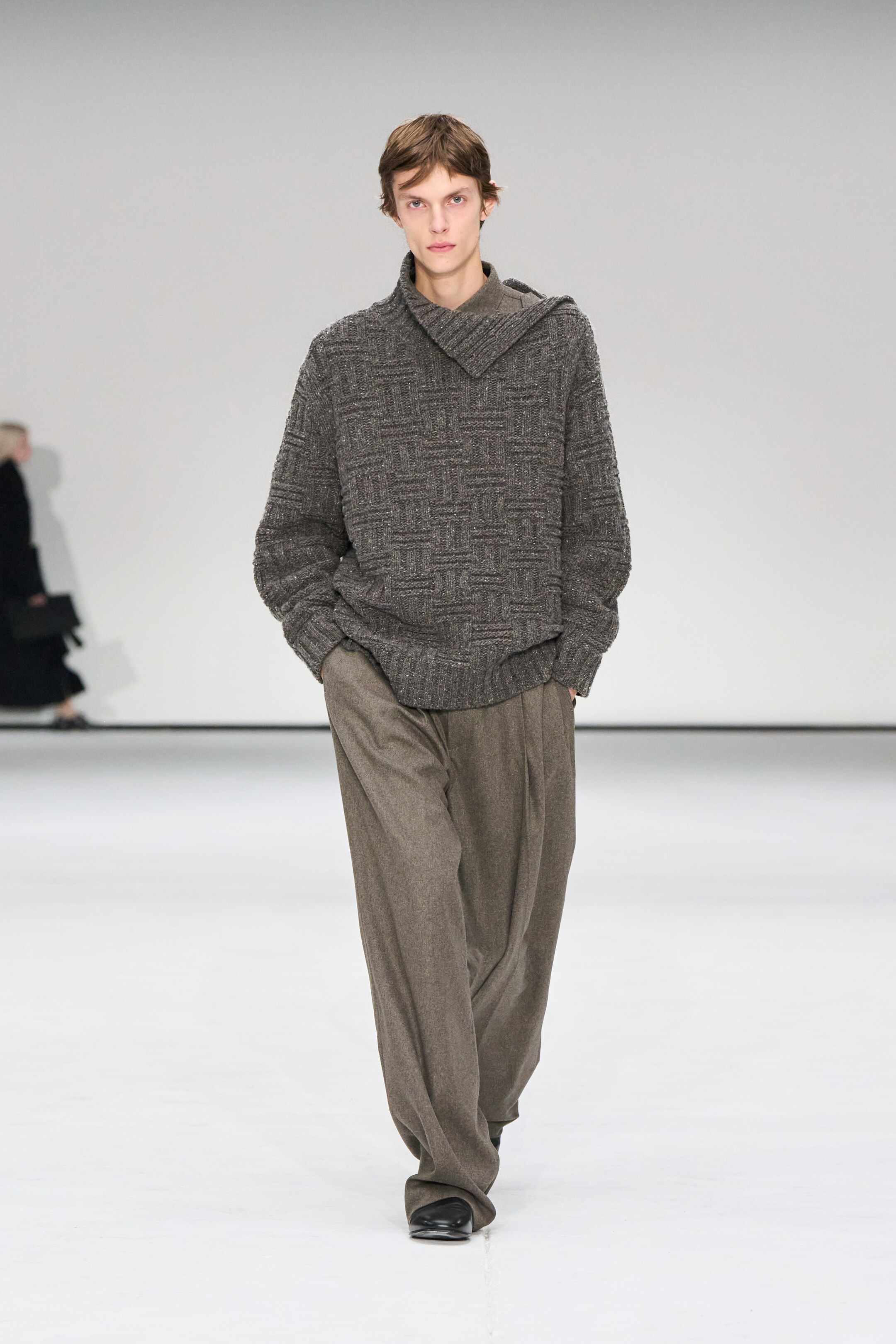 Größeres Bild anzeigen: Ein Model präsentiert einen dunkelgrauen, grob gestrickten Pullover und braune Faltenhosen von vorne. Der Pullover weist eine Korbgeflechtstruktur und einen gerippten Trichterkragen auf, der auf der linken Seite geknöpft ist, während die Hosen eine gerade, lockere Passform mit sichtbaren Gürtelschlaufen und Seitentaschen zeigen.