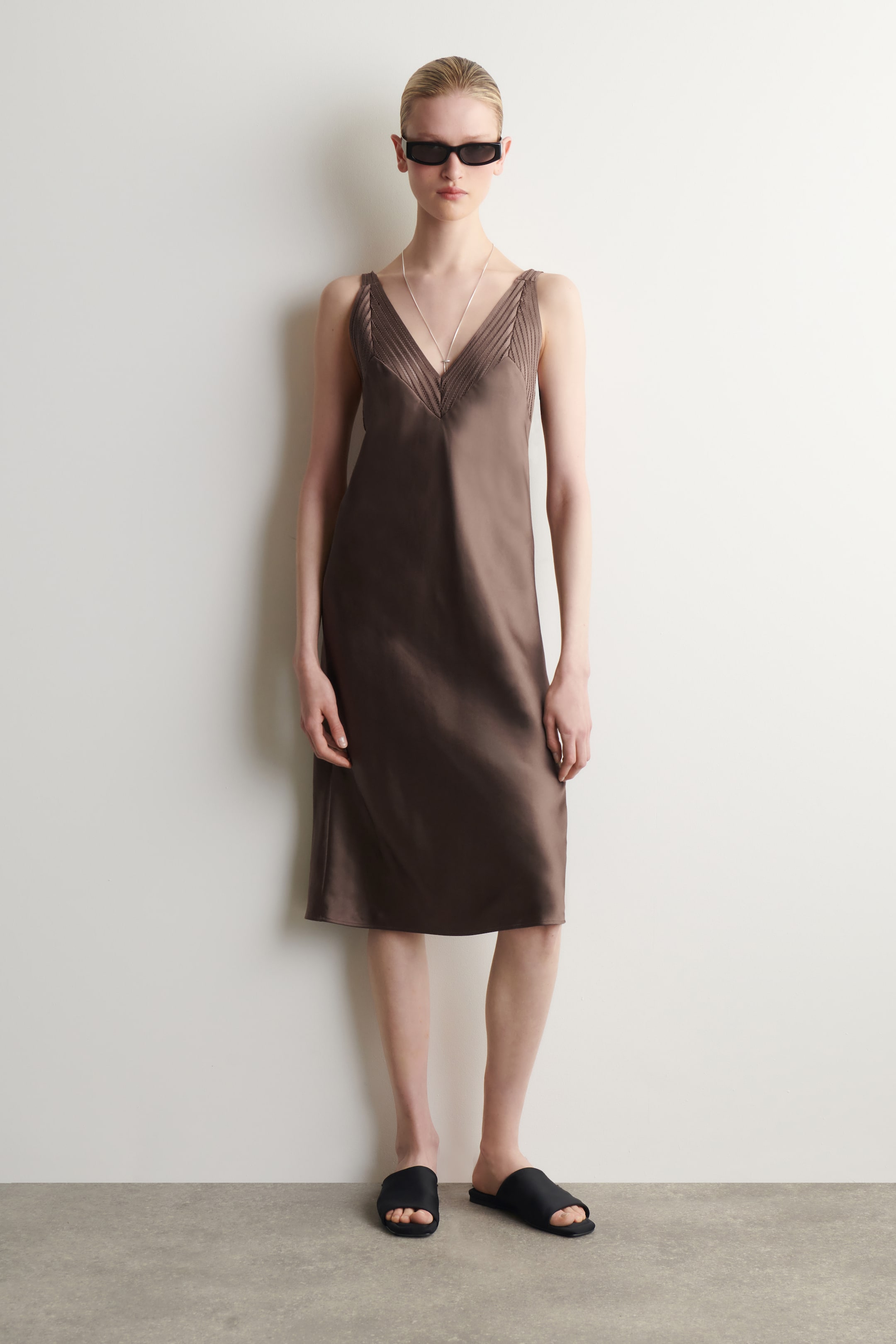Agrandir l'image: Une robe midi en satin marron est portée, avec un décolleté en V et des détails plissés sur les larges bretelles et le haut du corsage.
