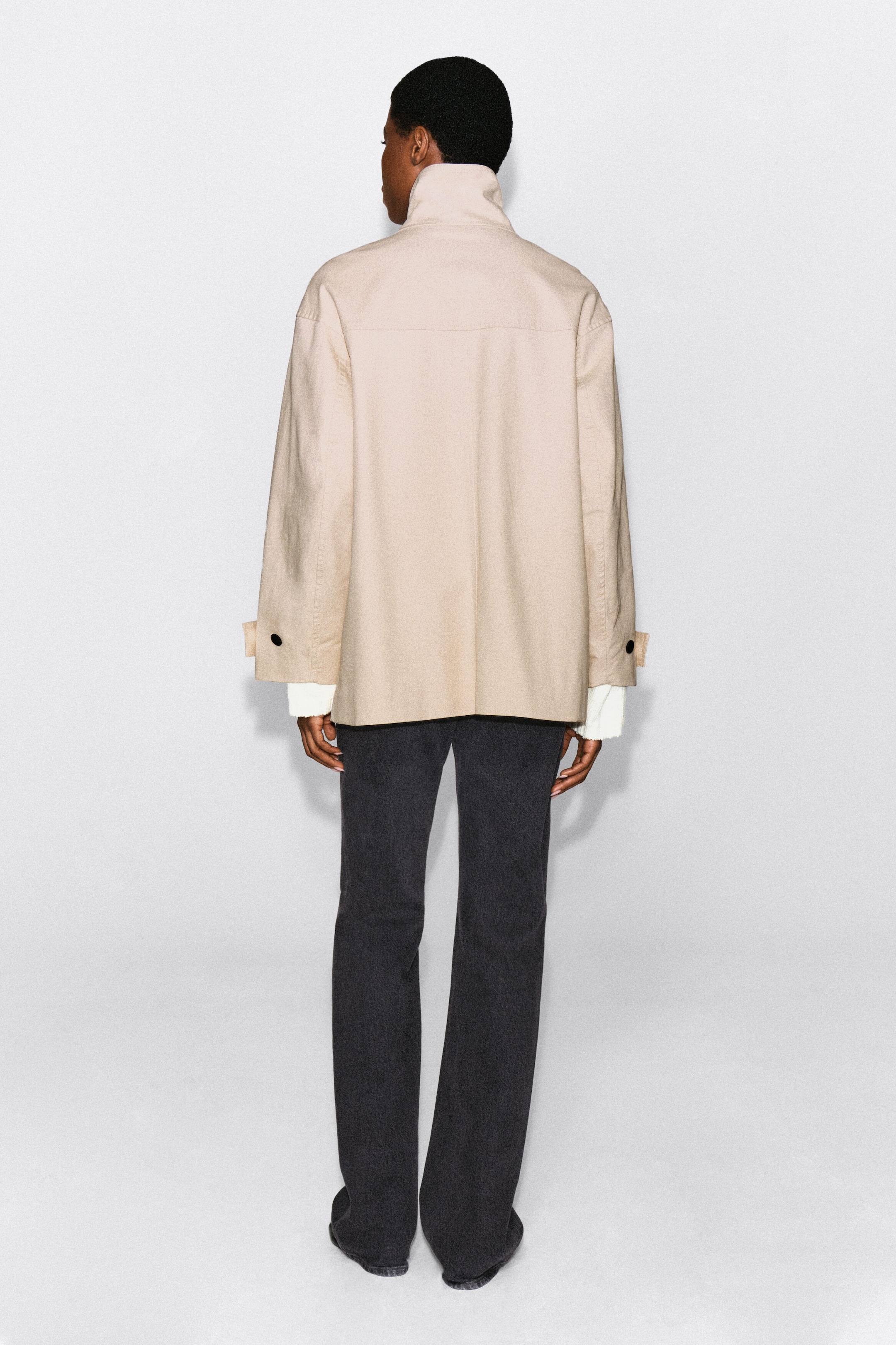 A beige cotton twill funnel-neck jacket is being worn by the model, displaying its back profile with a high collar, a horizontal back yoke, and buttoned cuff tabs. The jacket is layered over a white knit top and styled with dark grey trousers. A beige cotton twill funnel-neck jacket is being worn by the model, displaying its back profile with a high collar, a horizontal back yoke, and buttoned cuff tabs. The jacket is layered over a white knit top and styled with dark grey trousers.
