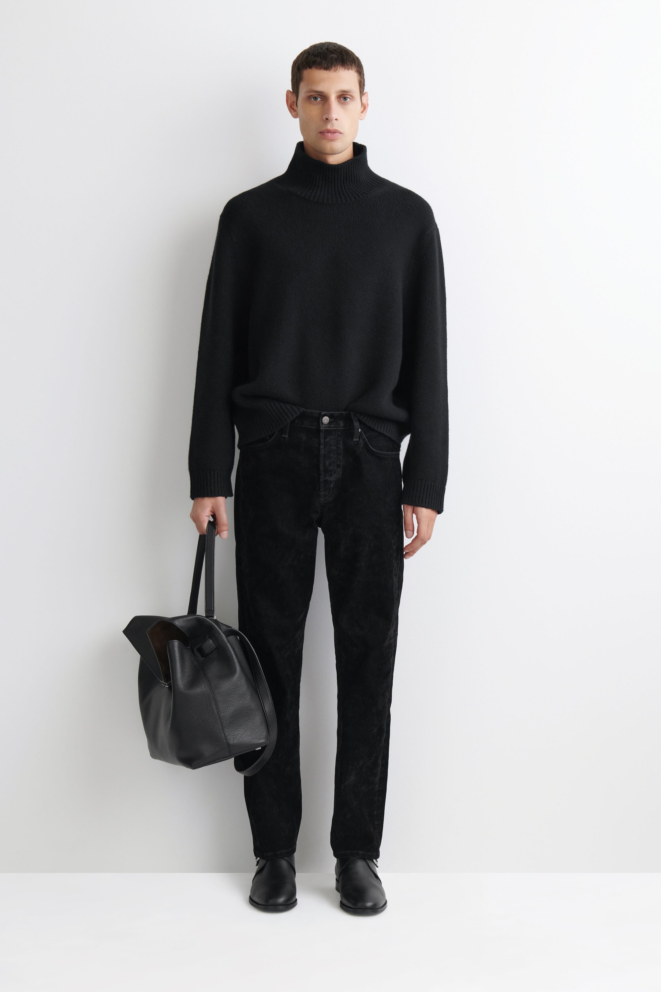 Con vista frontal, los Signature Flocked Straight-Leg Jeans presentan una silueta estructurada de pierna recta con un acabado flocado negro mate y texturizado. Los jeans se asientan a la cintura con un cierre clásico de botón, combinados con un jersey negro de cuello alto y zapatos de piel negros para destacar su bajo limpio y su caída relajada.