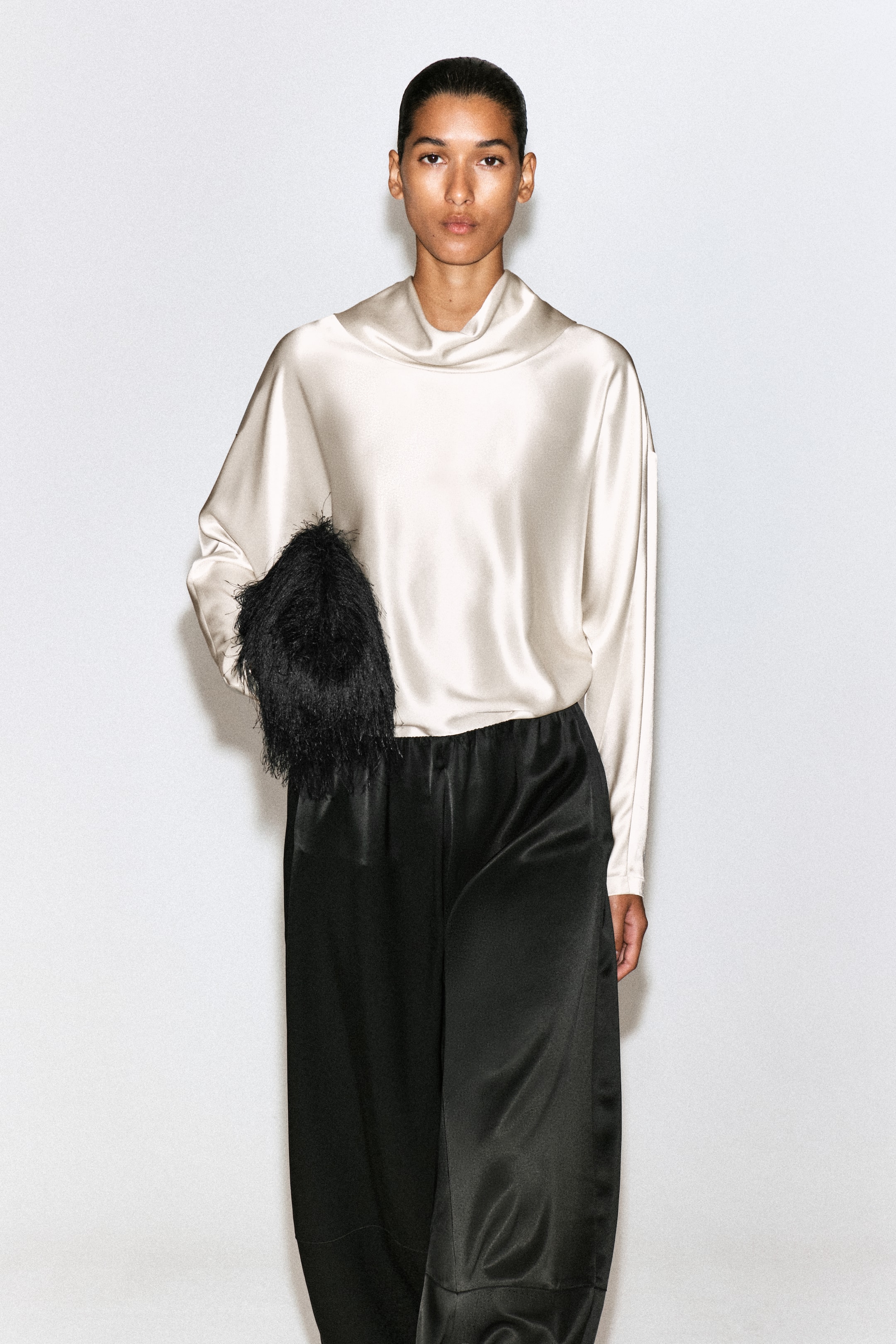 View larger image: A model facing front is wearing a champagne-coloured cowl-neck satin blouse. The loose-fitting blouse features a draped cowl neckline, dropped shoulders and long sleeves, and is styled with black trousers.