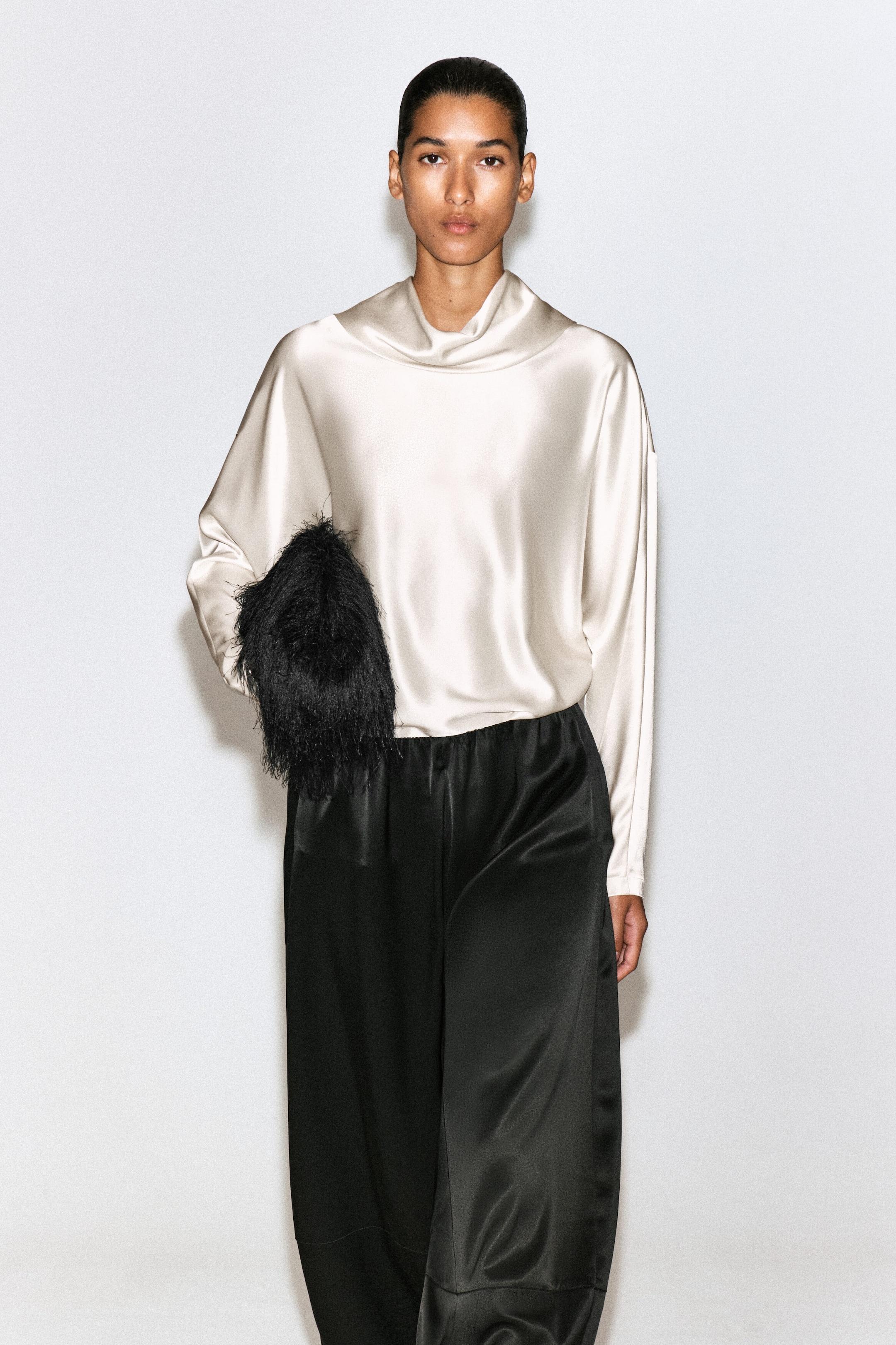A model facing forward is wearing a champagne-colored cowl-neck satin blouse. The loose-fitting blouse features a draped cowl neckline, dropped shoulders, and long sleeves, and is styled with black trousers.