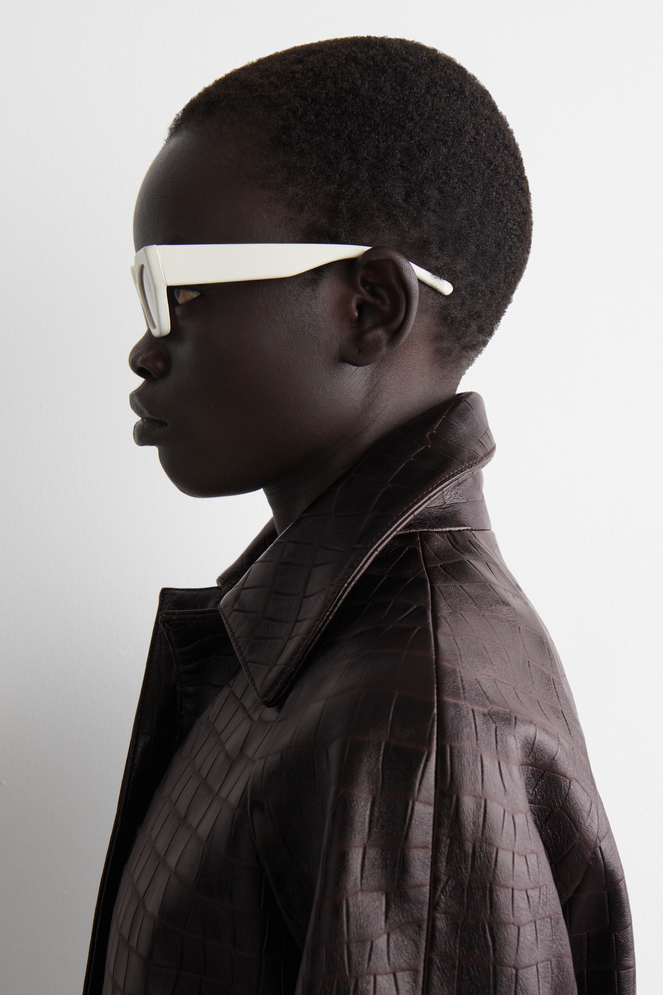 View larger image: A dark brown jacket, featuring a raised embossed crocodile pattern, is being worn. White, thick-framed sunglasses are also being worn, facing left.