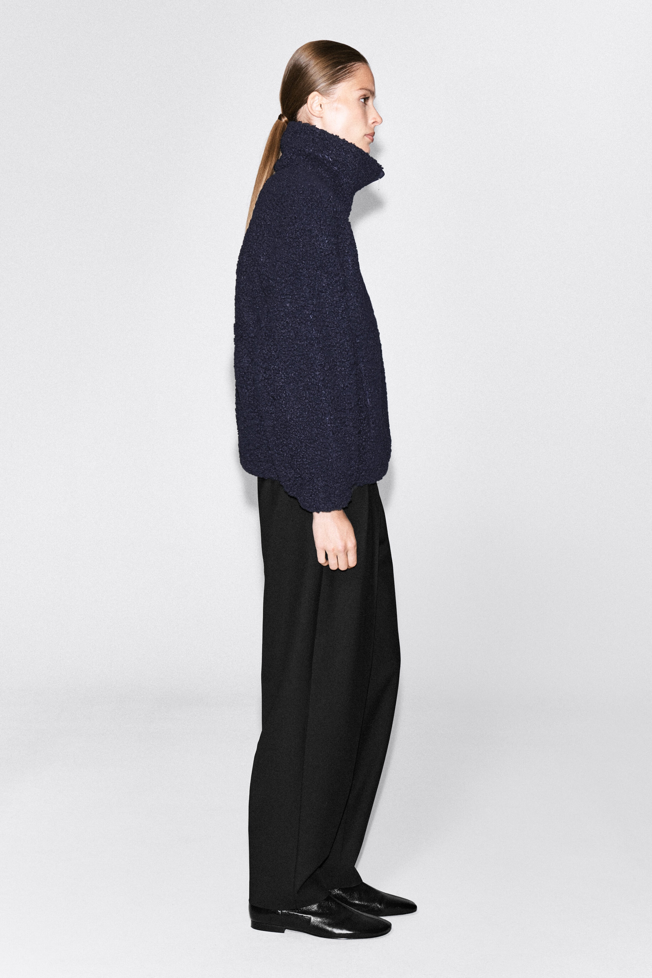 A dark navy, textured teddy sweater with a high funnel neck is being worn by the model, who is shown in side profile. The relaxed-fit pullover is paired with wide-leg black trousers and black leather shoes. A dark navy, textured teddy sweater with a high funnel neck is being worn by the model, who is shown in side profile. The relaxed-fit pullover is paired with wide-leg black trousers and black leather shoes.
