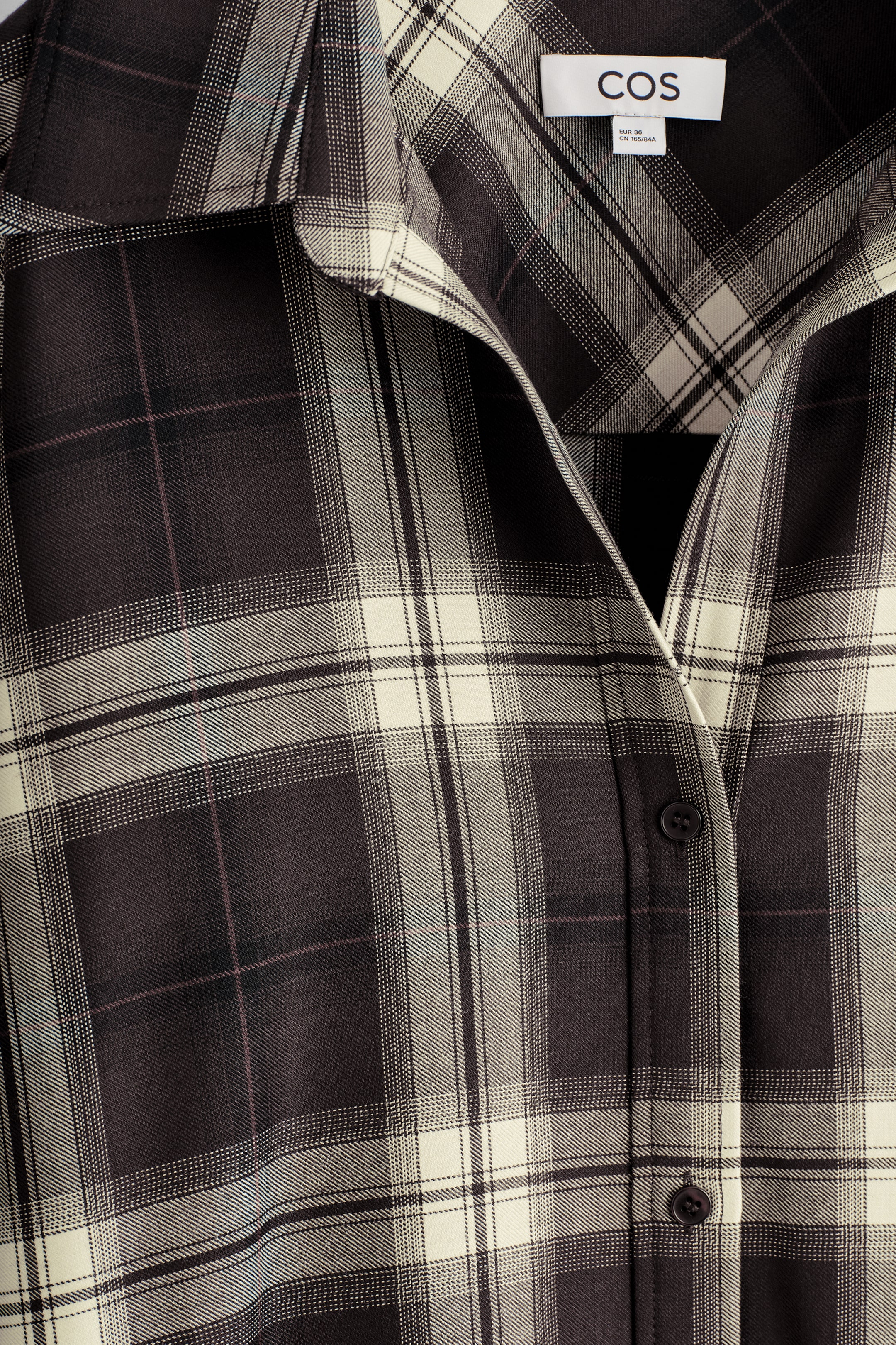 Visualizza immagine più grande: La PARTE ANTERIORE di un abito-camicia è mostrata, caratterizzata da un motivo a quadri marrone scuro e crema con sottili linee leggere. Presenta un colletto aperto che si piega all'indietro, formando un'apertura a V sopra un'abbottonatura visibile.