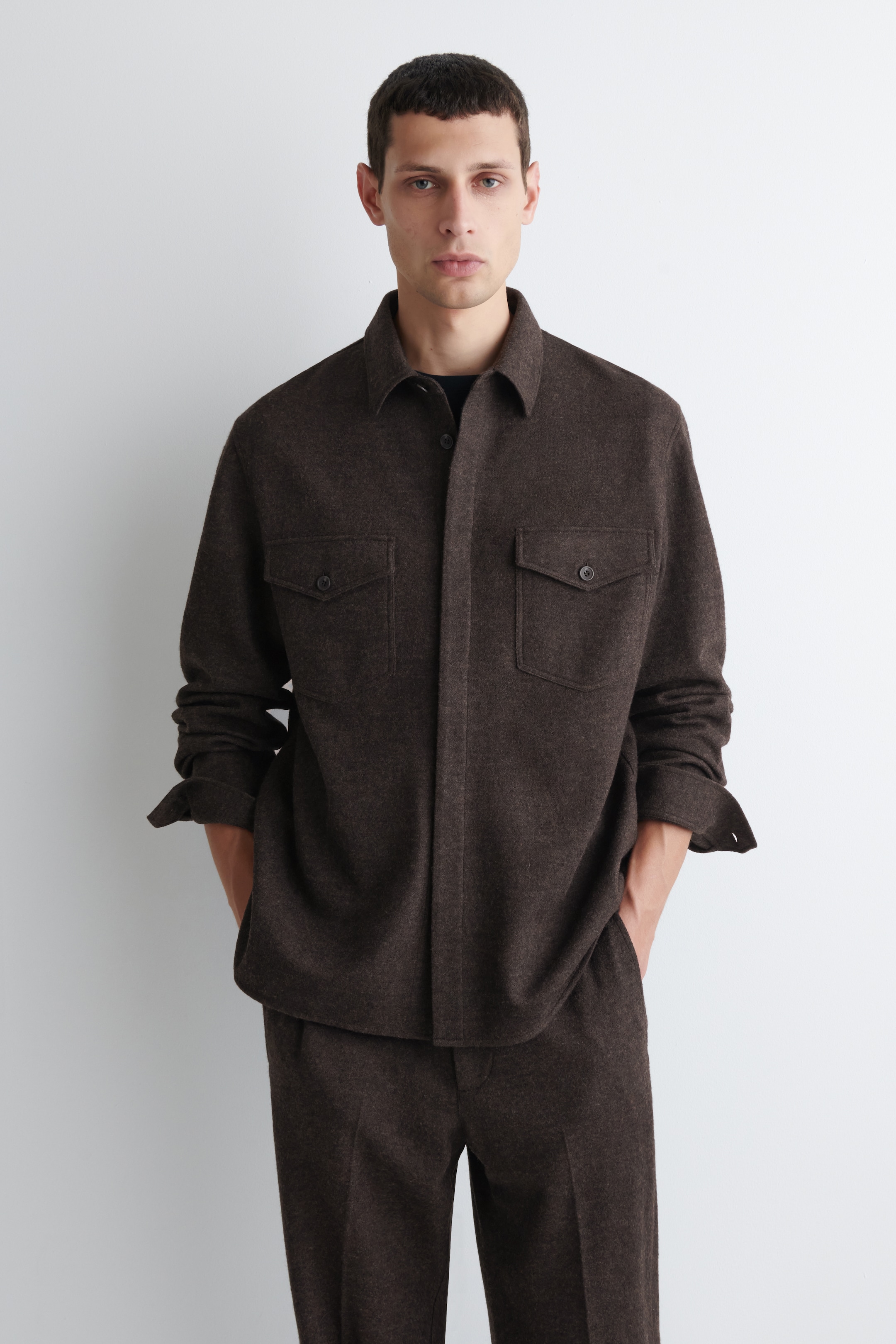 Agrandir l'image: Une veste-chemise marron foncé est présentée, avec un col standard, une patte de boutonnage à l'avant et deux poches poitrine à rabat avec fermetures à boutons. Les manches de la veste sont portées avec les poignets légèrement retroussés, et elle est superposée à un sous-vêtement foncé. Un pantalon marron foncé assorti complète l'ensemble décontracté, avec les mains reposant dans les poches latérales du pantalon, mettant en valeur l'ajustement et le drapé du vêtement.