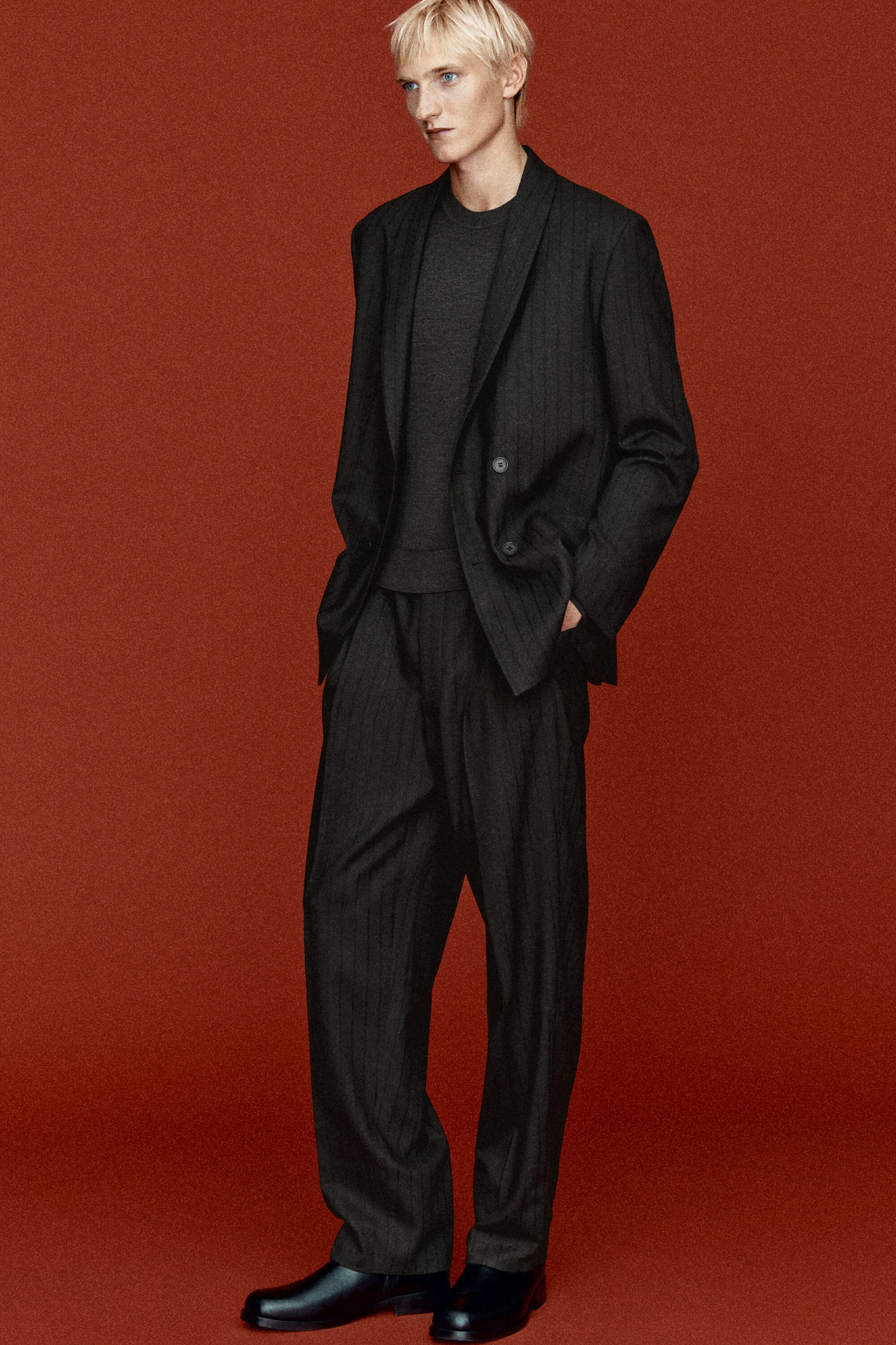 A dark gray pinstriped wool twill suit is shown in a front-facing view, featuring a relaxed-fit, double-breasted blazer with a shawl collar and matching relaxed, press-creased trousers. The blazer is worn open over a dark gray crewneck top, highlighting the suit’s modern tailoring. A dark gray pinstriped wool twill suit is shown in a front-facing view, featuring a relaxed-fit, double-breasted blazer with a shawl collar and matching relaxed, press-creased trousers. The blazer is worn open over a dark gray crewneck top, highlighting the suit’s modern tailoring.