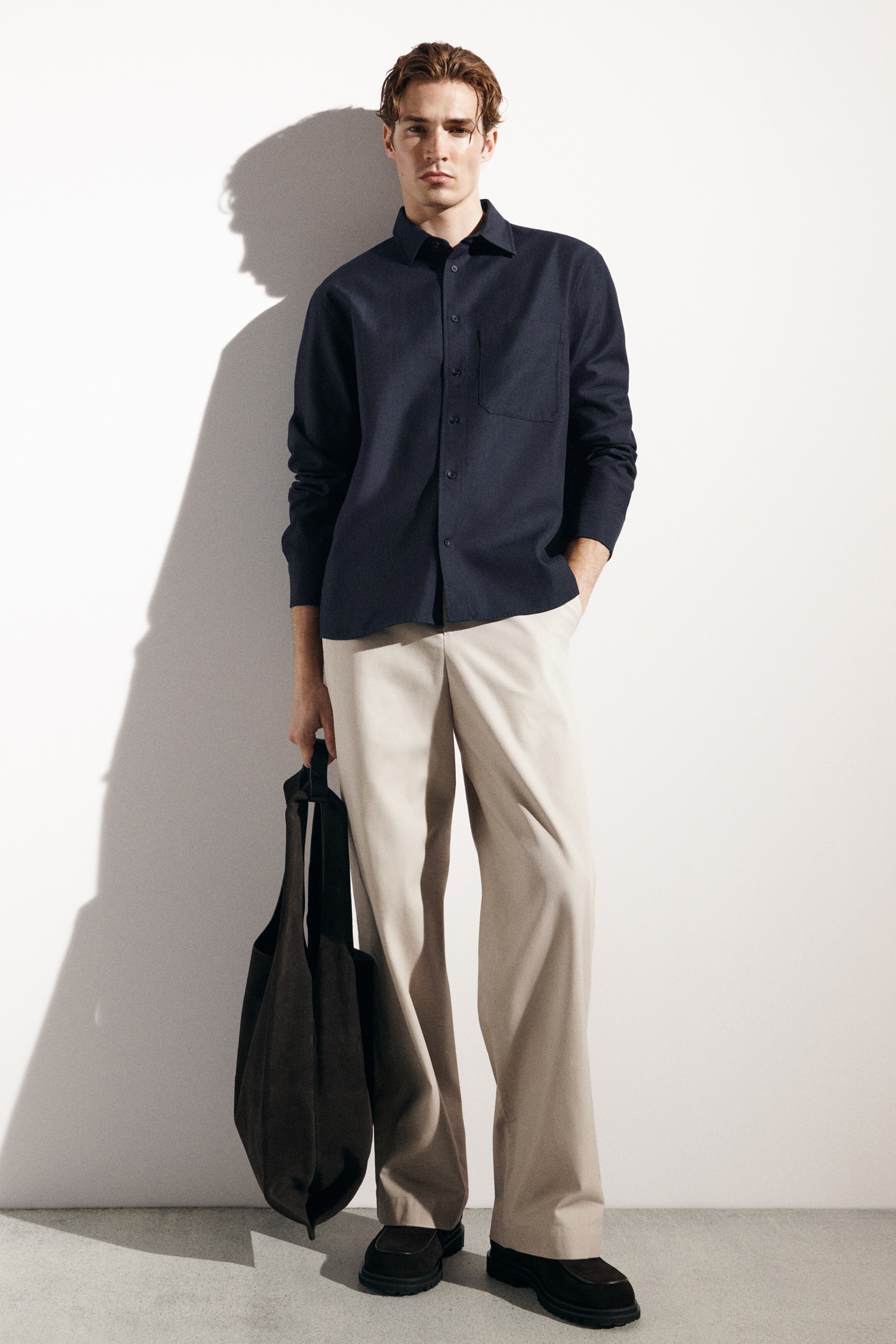 Größeres Bild anzeigen: Ein dunkelblaues Twill-Hemd mit spitzem Kragen, vollständiger Knopfleiste und einer einzelnen Brusttasche wird vorne getragen, in hellbeige, weit geschnittene Chinos mit sauberem Bund und Seitentaschen gesteckt. Eine dunkelbraune, lässige Wildleder-Tote-Bag mit breiten schwarzen Schulterriemen wird in der Hand gehalten.