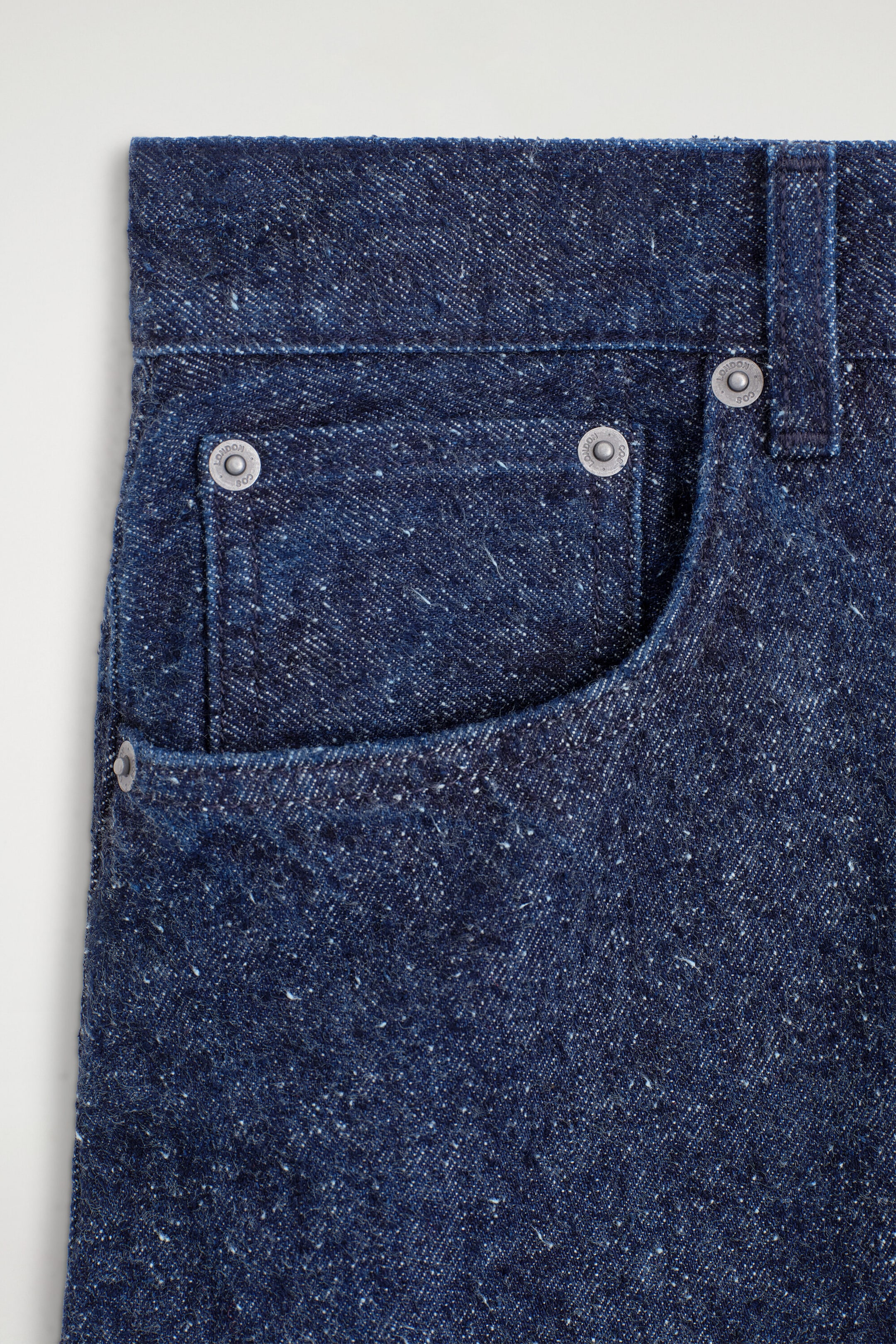 Dark blue denim jeans feature a front pocket with a curved opening and a smaller coin pocket. Silver-toned rivets reinforce the pocket corners, and the speckled fabric shows white flecks throughout. Dark blue denim jeans feature a front pocket with a curved opening and a smaller coin pocket. Silver-toned rivets reinforce the pocket corners, and the speckled fabric shows white flecks throughout.