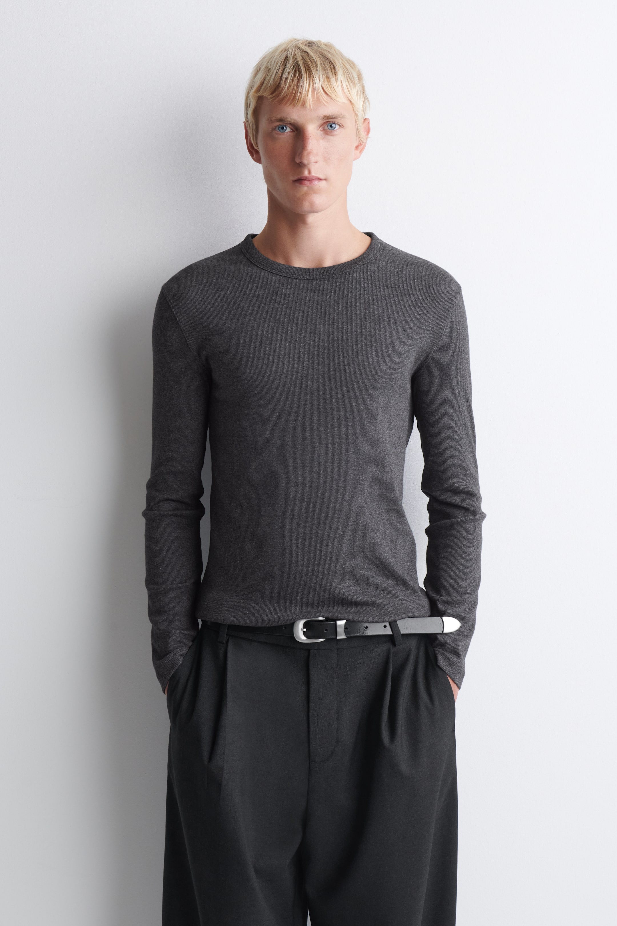 Ver imagen más grande: Una prenda de cuello redondo y manga larga en gris oscuro, vista de frente, que muestra una silueta entallada y una textura de punto fino. La prenda se lleva por dentro de unos pantalones oscuros de pinzas sujetos con un cinturón negro, resaltando su diseño limpio y ceñido.