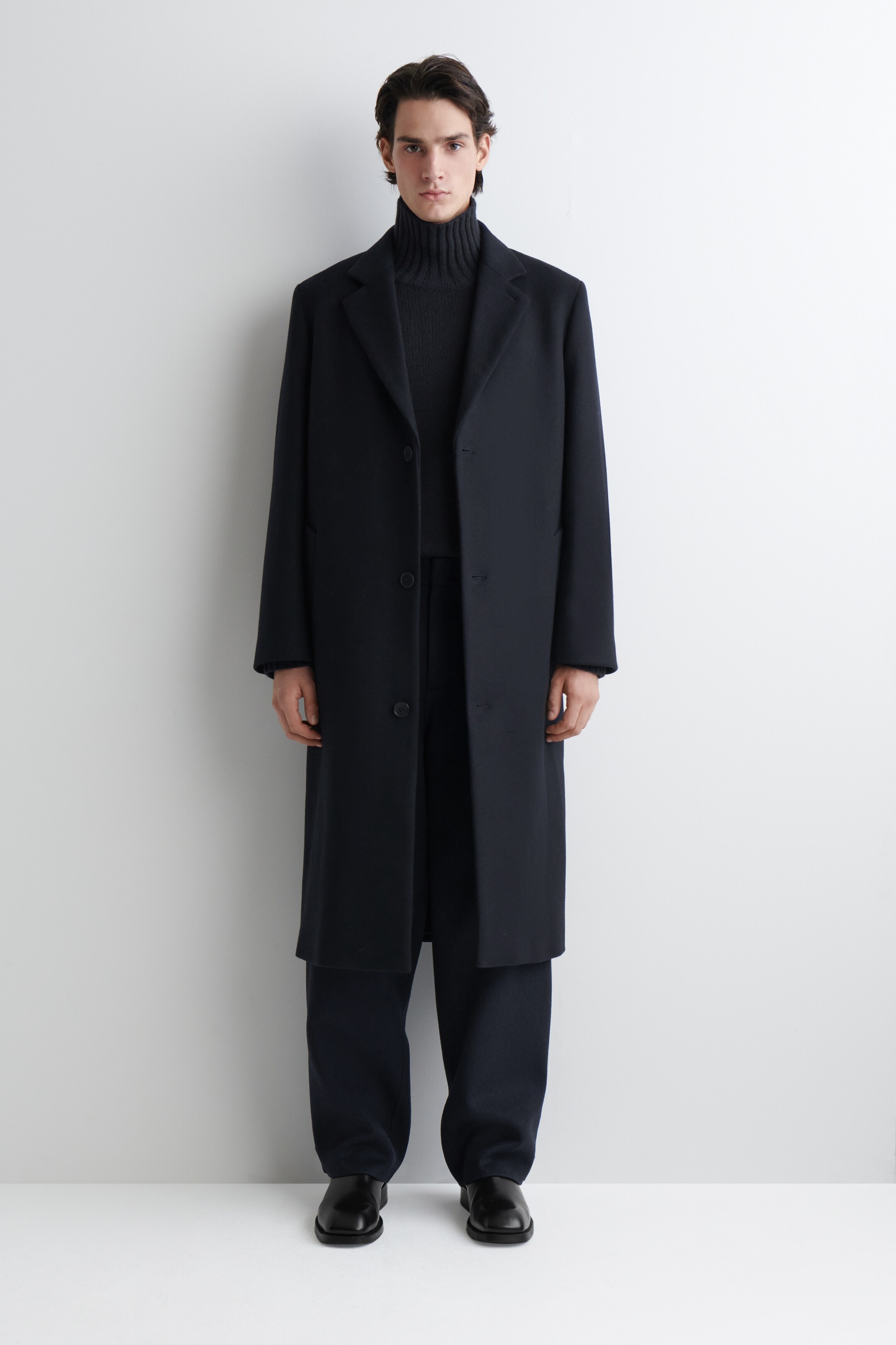 View larger image: A dark navy long coat is being presented from the front, featuring notch lapels and a single-breasted button closure. It is being worn over a dark ribbed turtleneck and dark trousers, with the coat falling to ankle length.