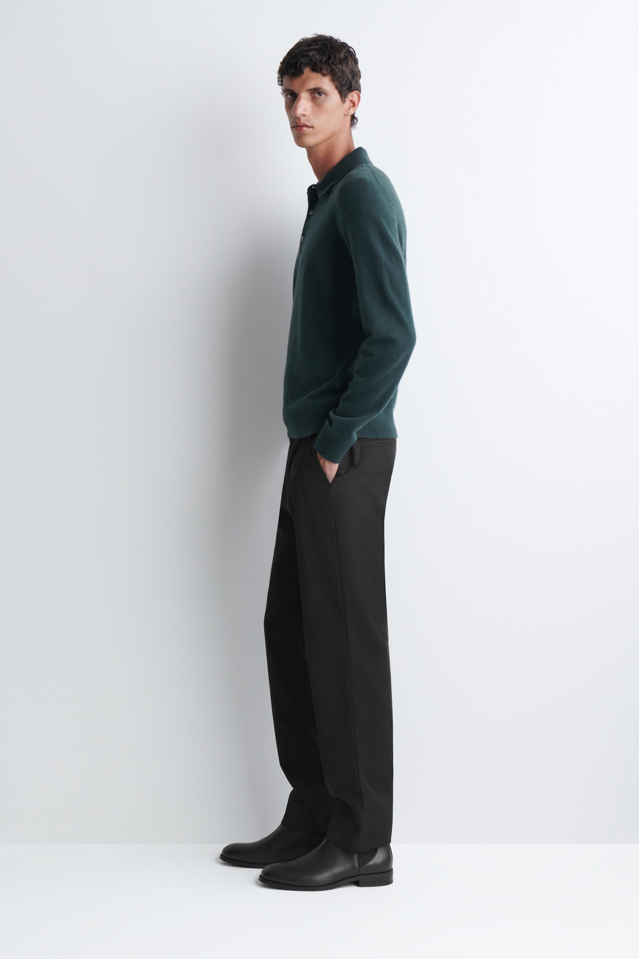 The dark green cashmere polo jumper, featuring a classic collar, a button placket, and long sleeves, is being worn by the model. The jumper is styled with black trousers and black leather boots as the model stands in profile.