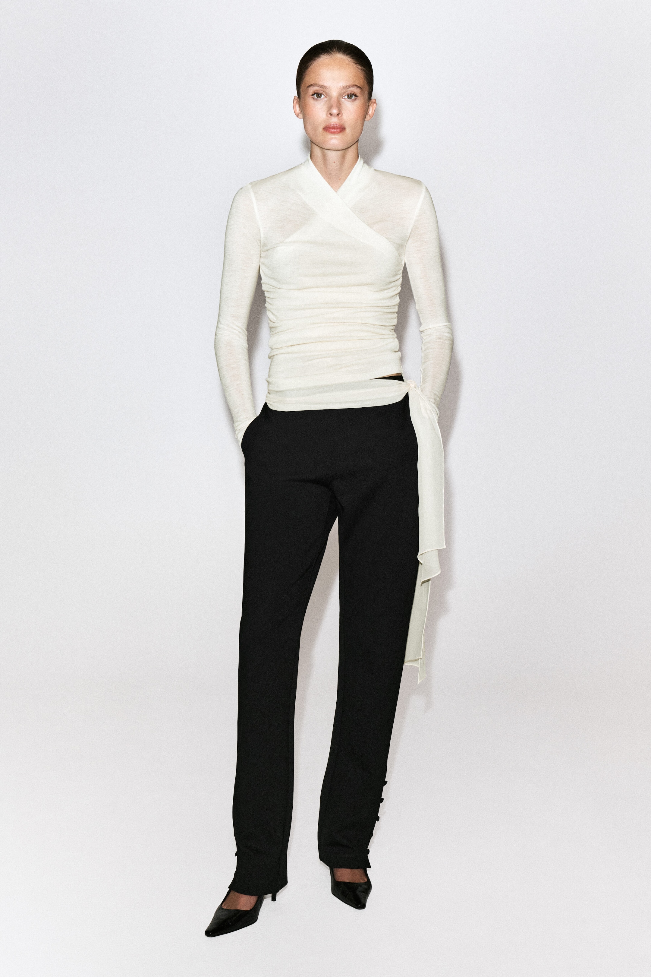 A cream-coloured, long-sleeved wrap top featuring a shawl collar, ruched detailing, and a long draped side panel is being worn by the model. The top is styled with black trousers and black pointed-toe heels. A cream-coloured, long-sleeved wrap top featuring a shawl collar, ruched detailing, and a long draped side panel is being worn by the model. The top is styled with black trousers and black pointed-toe heels.
