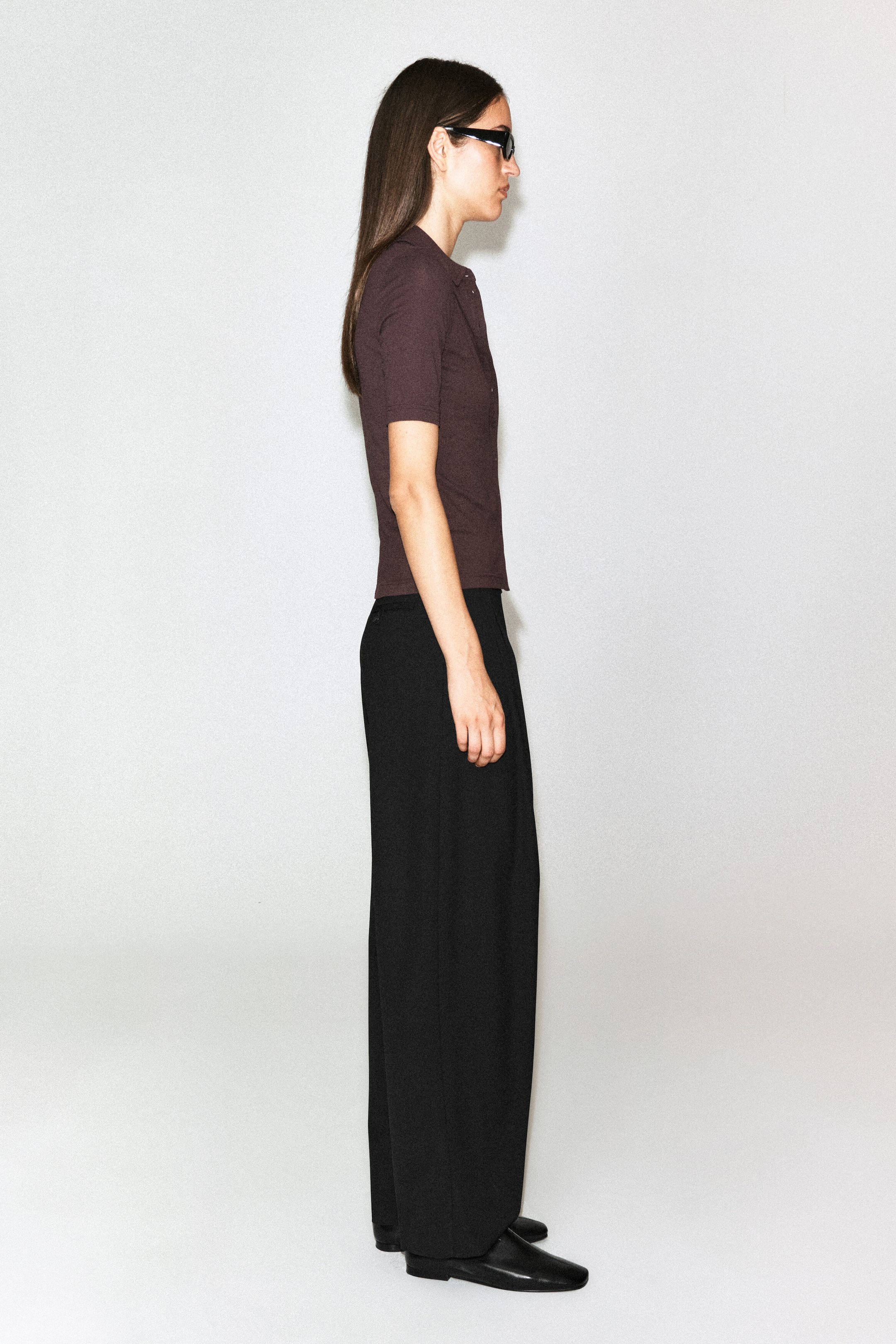 The sculpted cotton jersey shirt in dark brown is worn by the model, highlighting its slim fit, short sleeves, and classic collar from a side profile view. The shirt is styled with wide-leg black trousers and black sunglasses. The sculpted cotton jersey shirt in dark brown is worn by the model, highlighting its slim fit, short sleeves, and classic collar from a side profile view. The shirt is styled with wide-leg black trousers and black sunglasses.