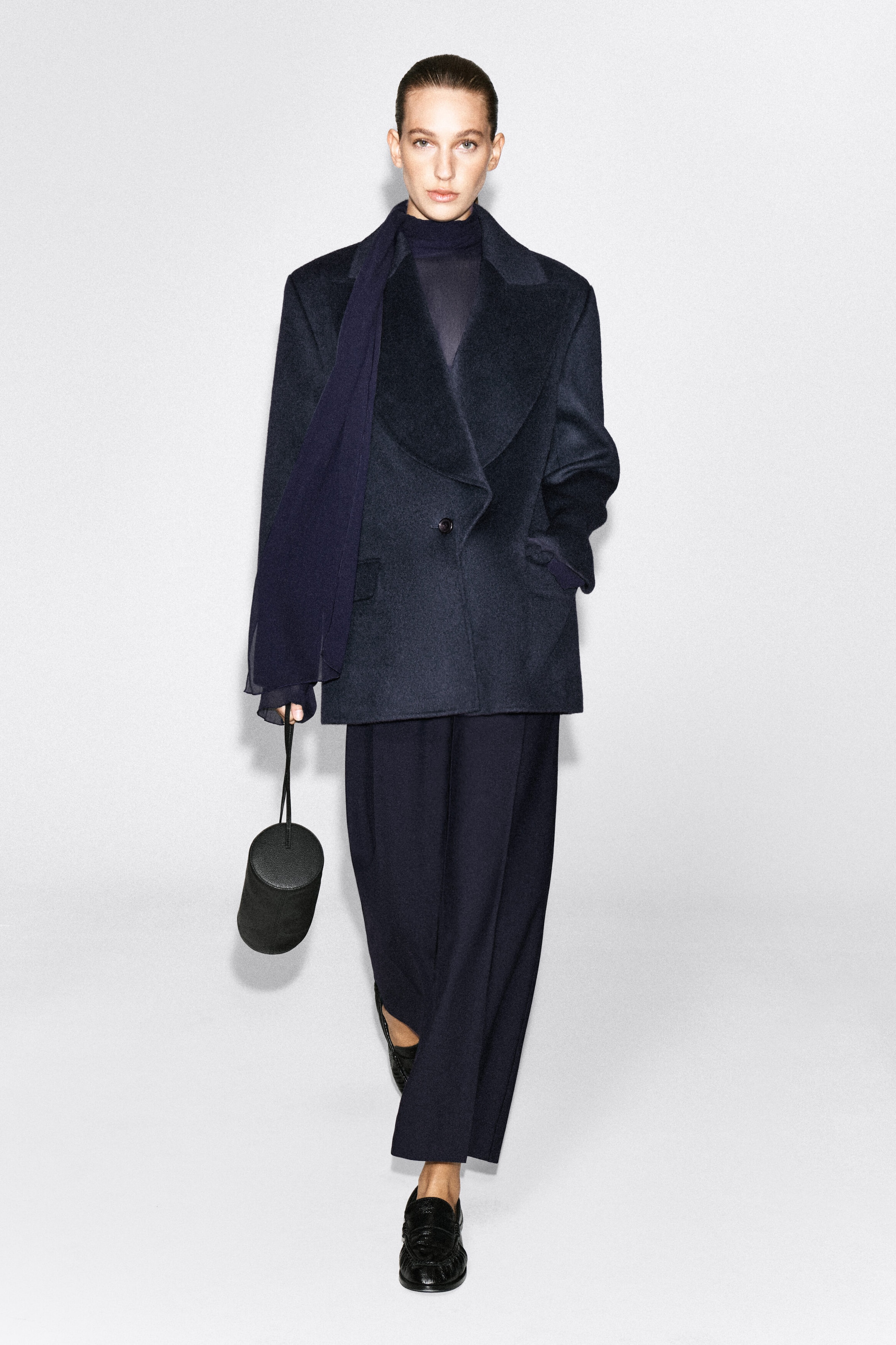A dark navy wool-blend blazer featuring exaggerated peak lapels and a single-button fastening is worn by the model. The blazer is styled over a dark high-neck top with matching wide-leg trousers, while the model holds a small cylindrical black bag in one hand.