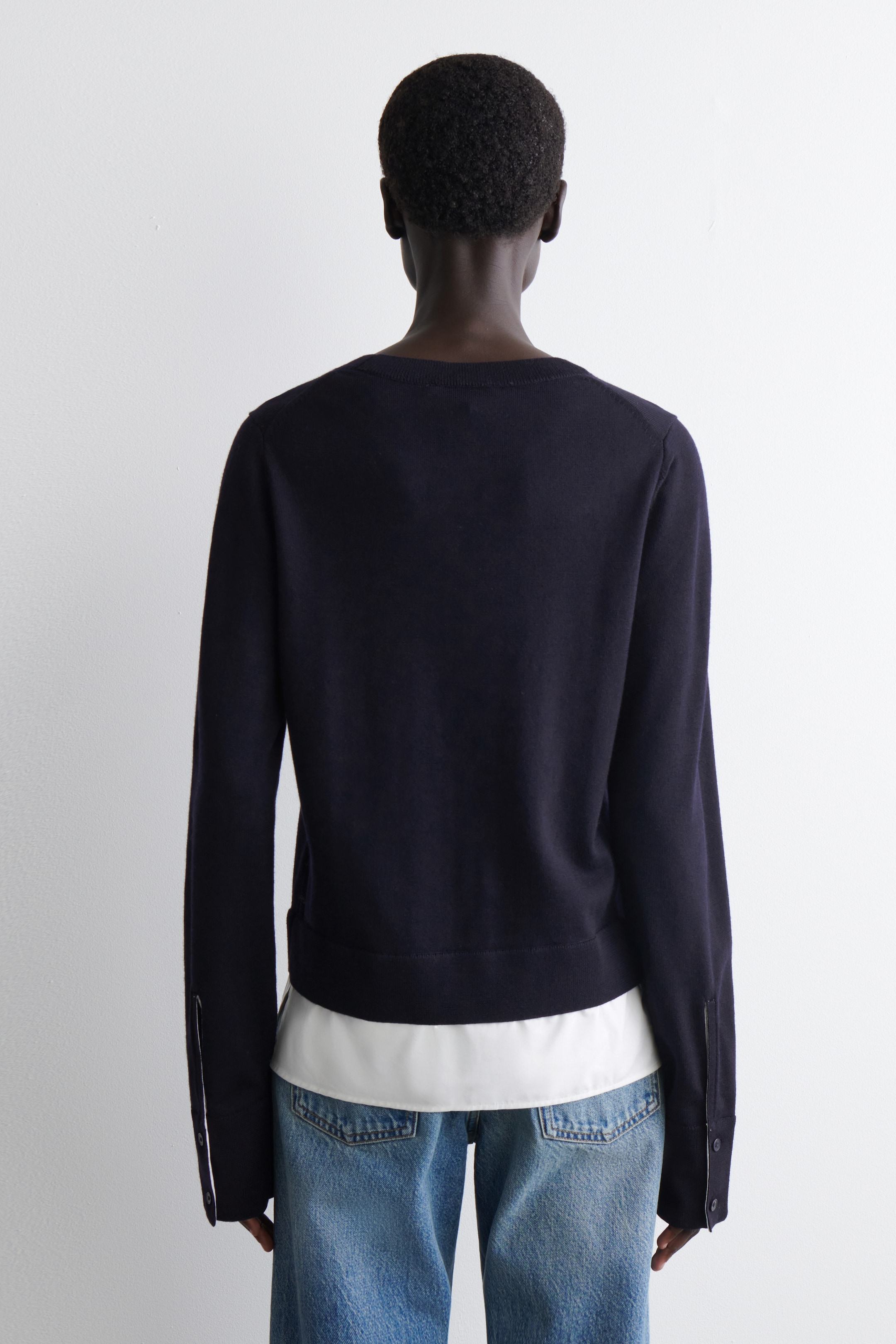 Visualizza immagine più grande: Un maglione a maglia blu scuro è mostrato dal retro, caratterizzato da uno scollo a girocollo e maniche lunghe con polsini a fessura con bottoni. Un indumento bianco è visibile, stratificato sotto l'orlo del maglione e che sbuca attraverso i polsini aperti.
