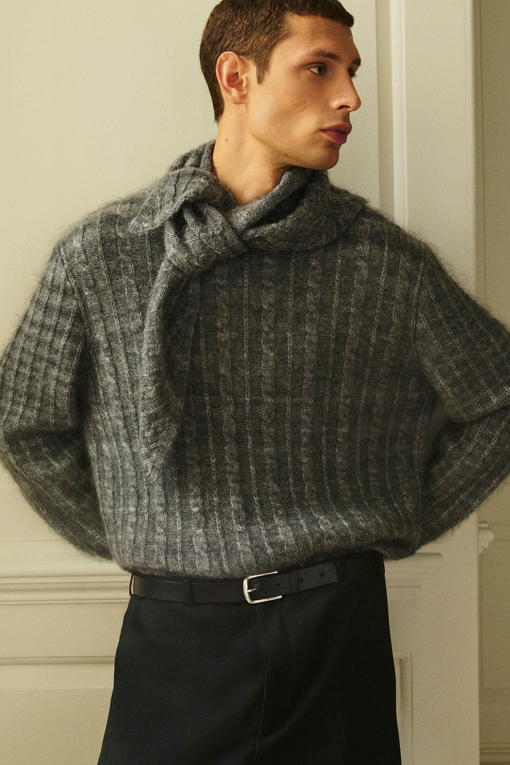 A grey cable-knit merino-mohair jumper is shown with a fuzzy, textured finish and vertical cable-stitch detailing in a front-facing view. The jumper features a matching knit scarf tied loosely around the neck and is worn tucked into dark trousers with a black leather belt. A grey cable-knit merino-mohair jumper is shown with a fuzzy, textured finish and vertical cable-stitch detailing in a front-facing view. The jumper features a matching knit scarf tied loosely around the neck and is worn tucked into dark trousers with a black leather belt.