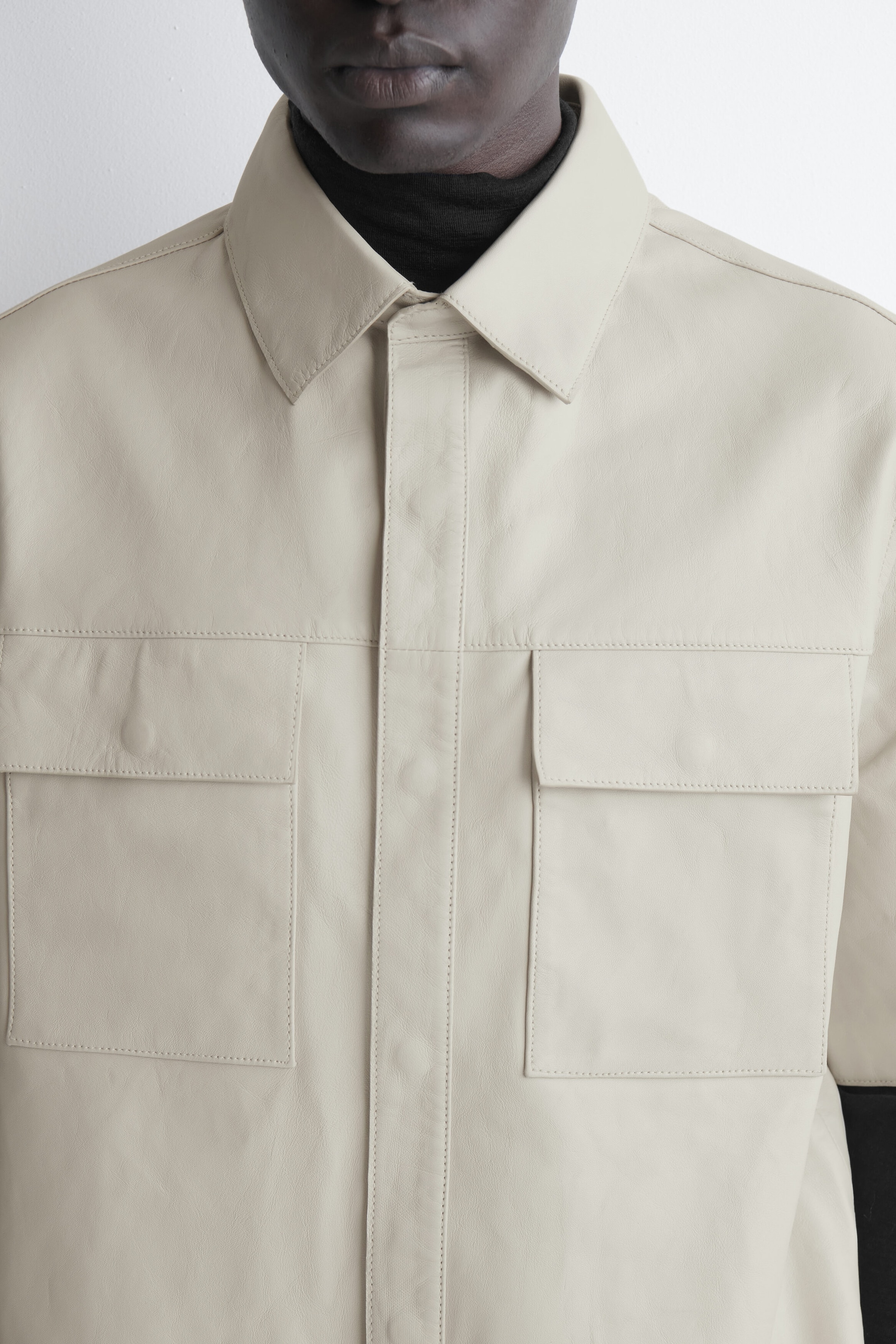Ein hellbeiges Lederhemd ist in einer nahen Frontansicht zu sehen und verfügt über einen spitzen Kragen, eine verdeckte Knopfleiste und zwei große Brust-Patchtaschen mit Patten. Das Hemd wird über einem schwarzen Rollkragenpullover getragen und hebt so sein strukturiertes Design sowie die sauberen, abgesteppten Nähte hervor.