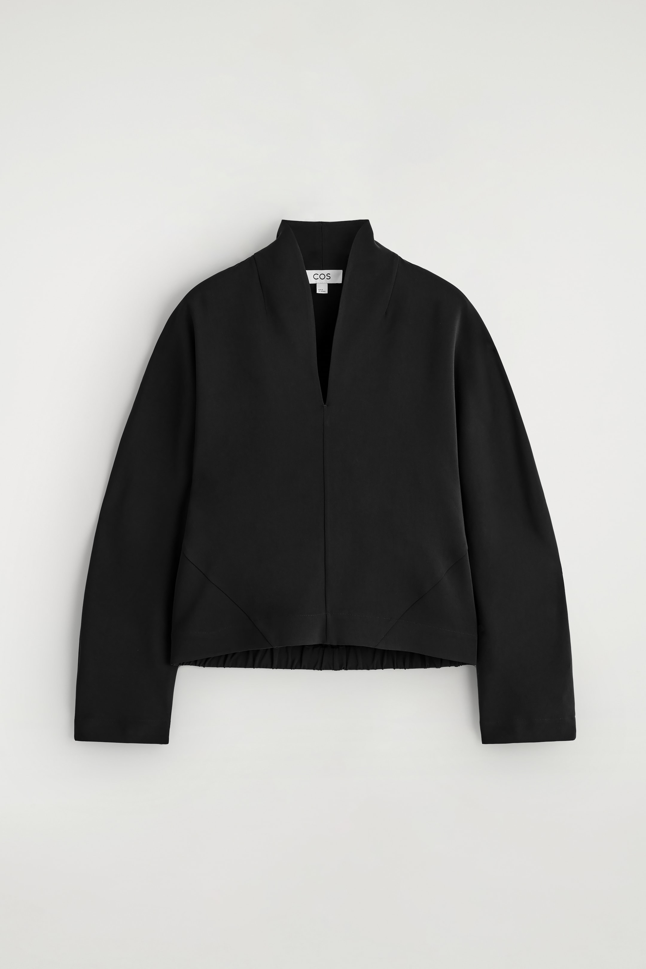 Visualizza immagine più grande: La parte anteriore di una blusa nera è visibile, caratterizzata da una vestibilità rilassata e un colletto alto che si apre in uno scollo a V scultoreo. È progettata con maniche lunghe e cuciture diagonali sopra un orlo elasticizzato.