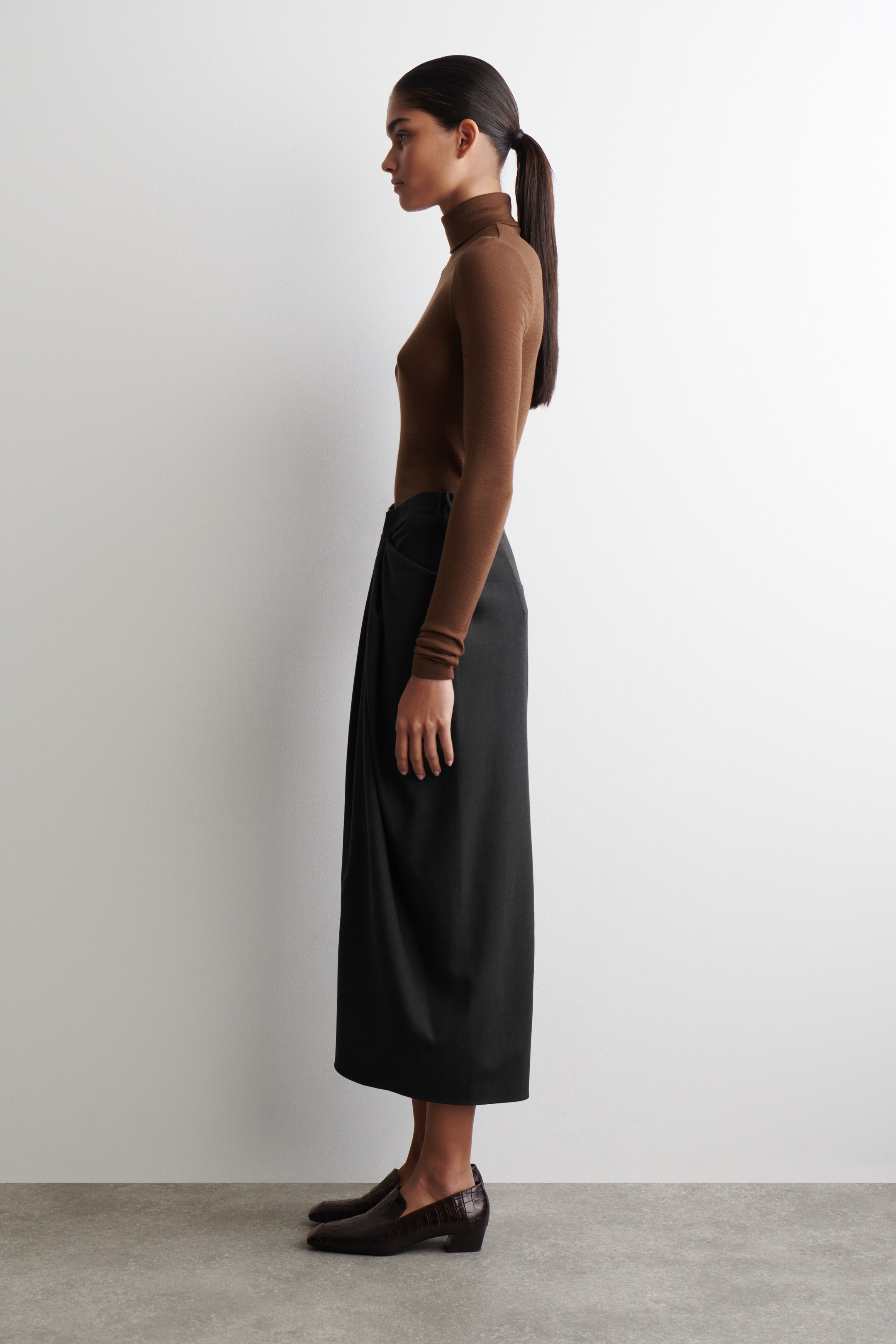Ver imagen más grande: Se lleva puesto un top de cuello alto marrón de manga larga, metido en una falda midi negra que presenta un detalle de cintura drapeada, todo visto de perfil. Unos mocasines de tacón bajo texturizados en marrón oscuro completan el conjunto.