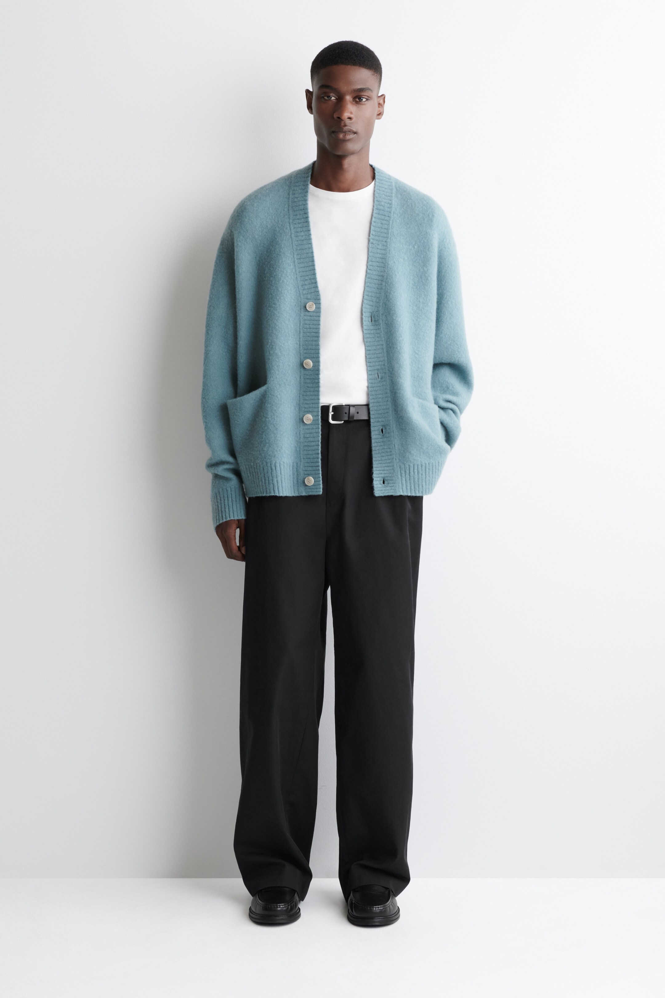 Ver imagen más grande: Una chaqueta de punto color verde azulado con una textura cepillada, un cuello en V acanalado y cierre de botones se lleva desabrochada sobre una camiseta blanca. La modelo está de pie con las manos en los bolsillos de parche frontales de la chaqueta.