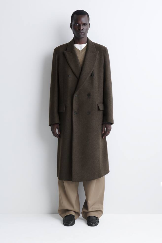 A double-breasted wool-blend long coat in dark olive brown is shown from the front, featuring wide notch lapels, a six-button front fastening, and side flap pockets. The structured, long-line coat is layered over a brown V-neck knit sweater and beige wide-leg trousers. - 1