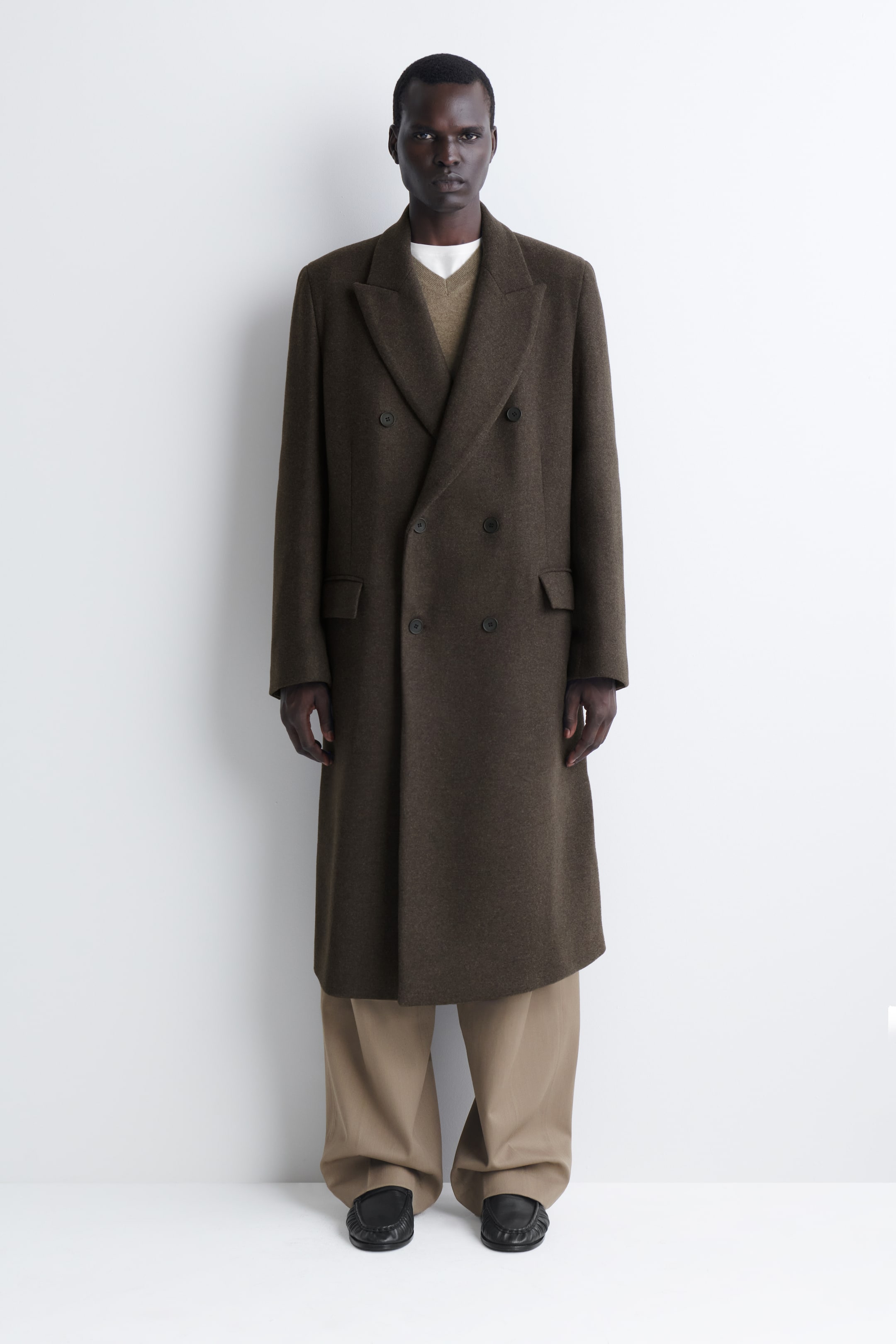 Agrandir l'image: Un long manteau en mélange de laine, à double boutonnage, de couleur brun olive foncé, est présenté de face, avec de larges revers à cran, une fermeture frontale à six boutons et des poches latérales à rabat. Ce manteau structuré à la ligne allongée est porté sur un pull en maille brun à col en V et un pantalon beige à jambes larges.