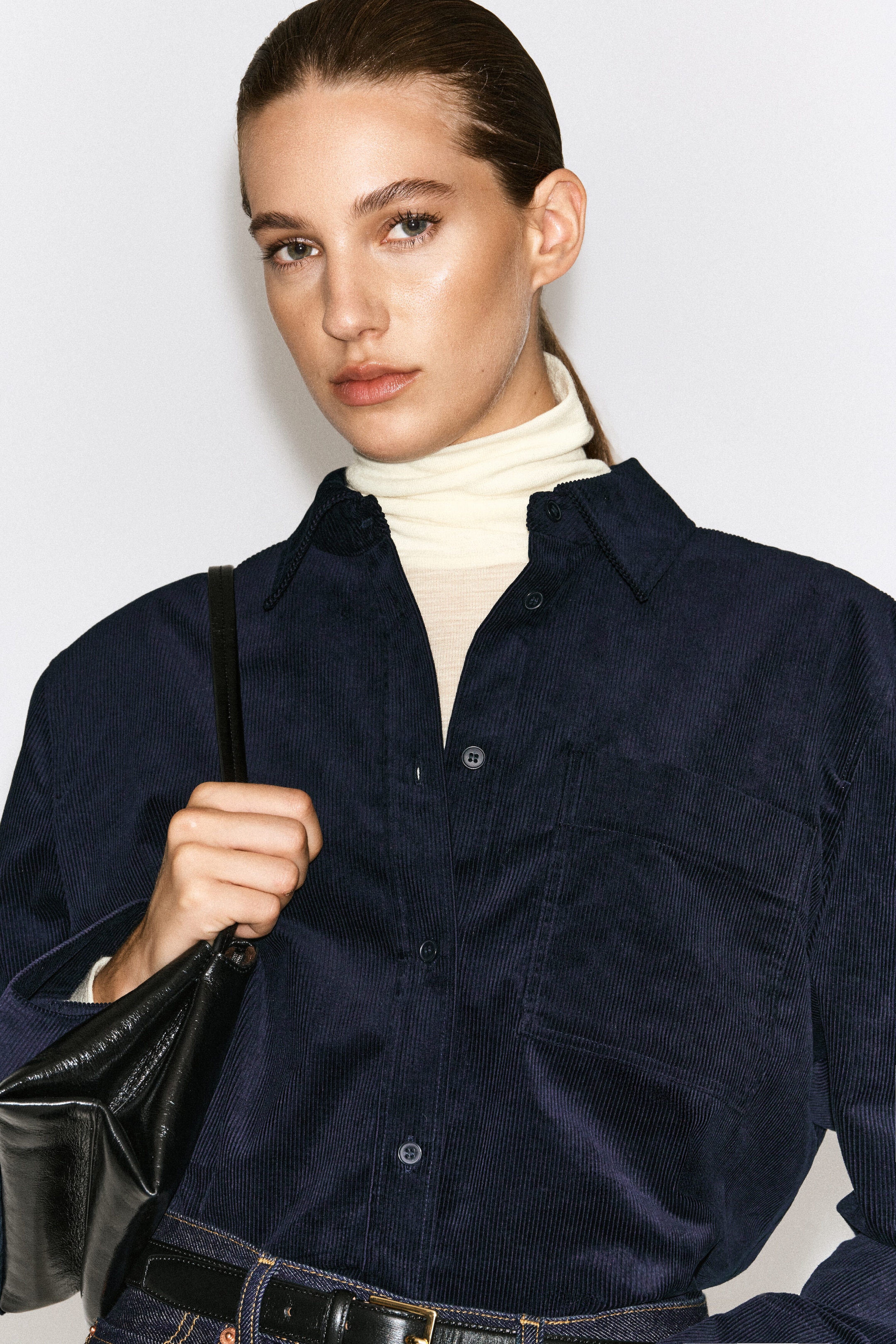 A dark navy blue cotton corduroy shirt is worn by the model, layered over a cream-colored turtleneck top. The shirt features a ribbed texture, a classic collar, and a front button closure, and is tucked into belted denim trousers while the model holds a black leather bag. A dark navy blue cotton corduroy shirt is worn by the model, layered over a cream-colored turtleneck top. The shirt features a ribbed texture, a classic collar, and a front button closure, and is tucked into belted denim trousers while the model holds a black leather bag.