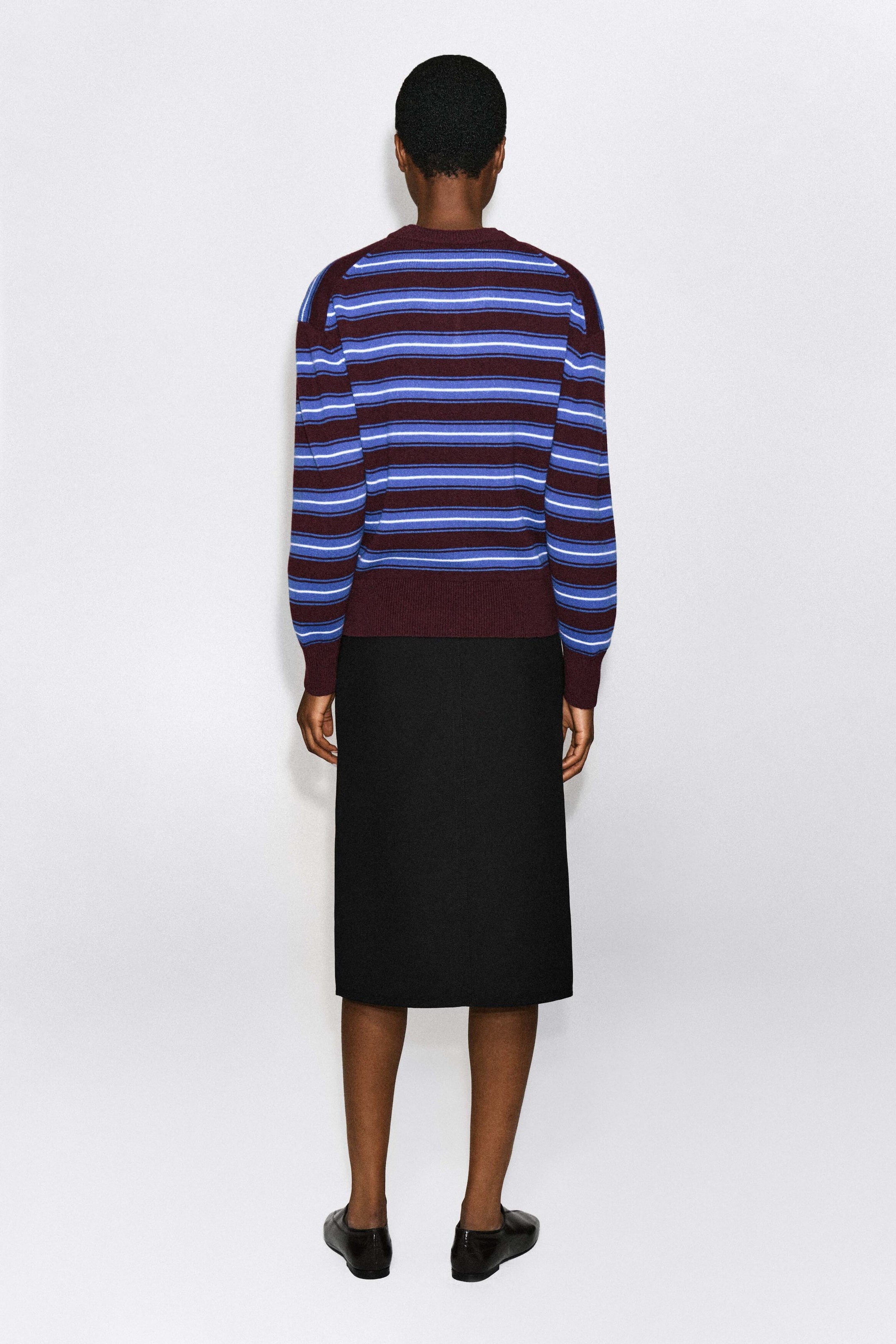 A merino wool sweater featuring horizontal stripes in burgundy, blue, and white is being worn by the model, shown from the back. The sweater is styled with a black midi skirt and black leather flat shoes. A merino wool sweater featuring horizontal stripes in burgundy, blue, and white is being worn by the model, shown from the back. The sweater is styled with a black midi skirt and black leather flat shoes.