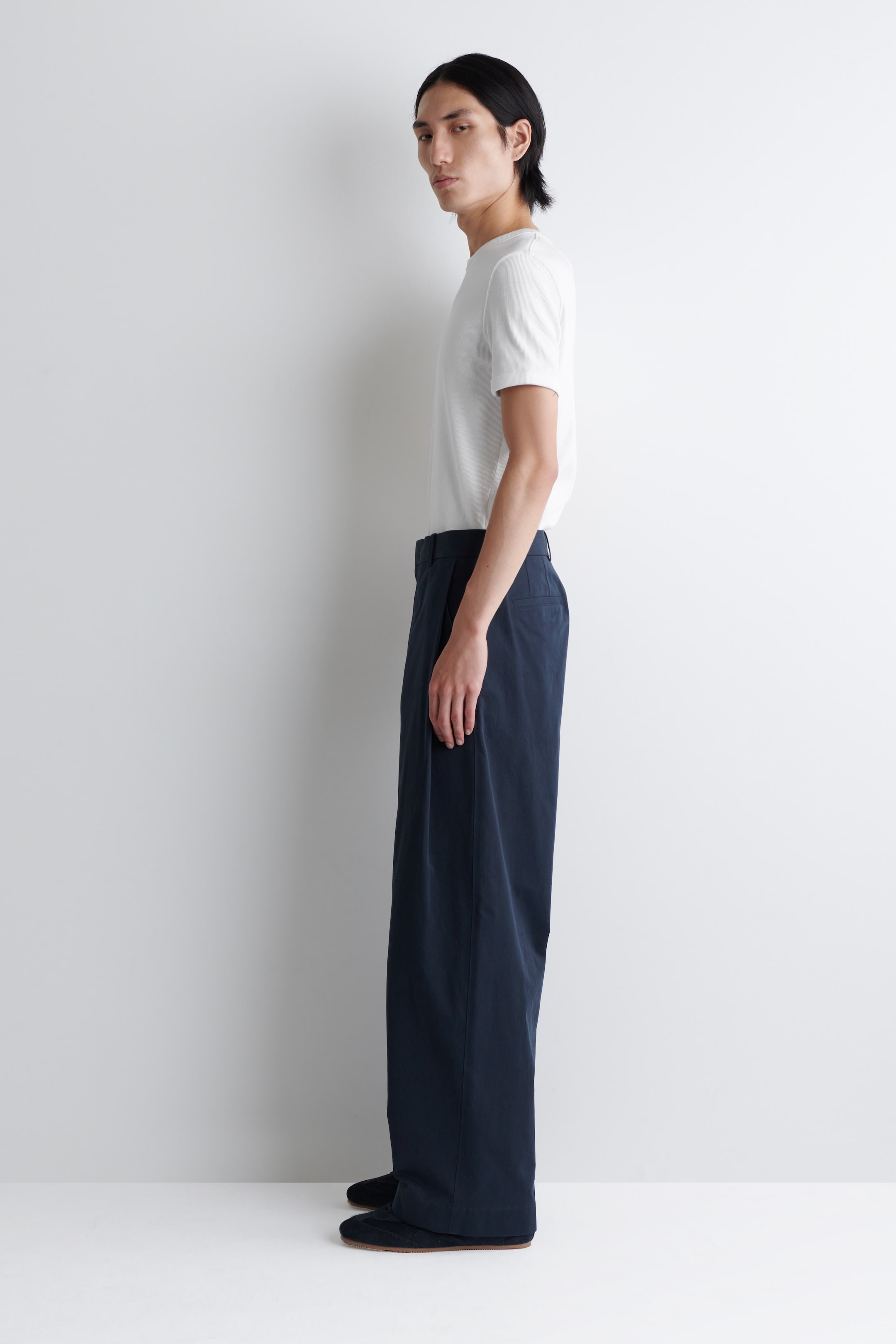 Visualizza immagine più grande: Una t-shirt bianca a maniche corte e pantaloni blu scuro a gamba larga vengono presentati di lato. La t-shirt, con scollo rotondo e vestibilità regolare, è indossata infilata nei pantaloni a vita alta, che mostrano una silhouette ampia e fluente e si accumulano leggermente sul pavimento.
