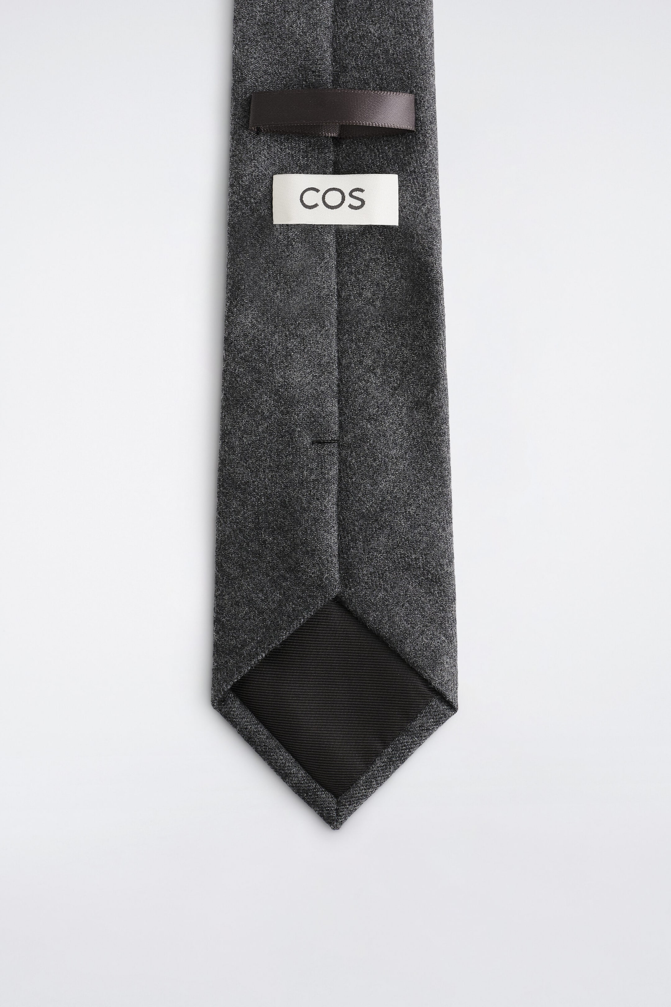 A dark grey textured tie is displayed underside up, featuring a dark brown satin keeper loop and a white label with the brand name "COS" printed in black. The pointed tip shows a black ribbed fabric lining. A dark grey textured tie is displayed underside up, featuring a dark brown satin keeper loop and a white label with the brand name "COS" printed in black. The pointed tip shows a black ribbed fabric lining.